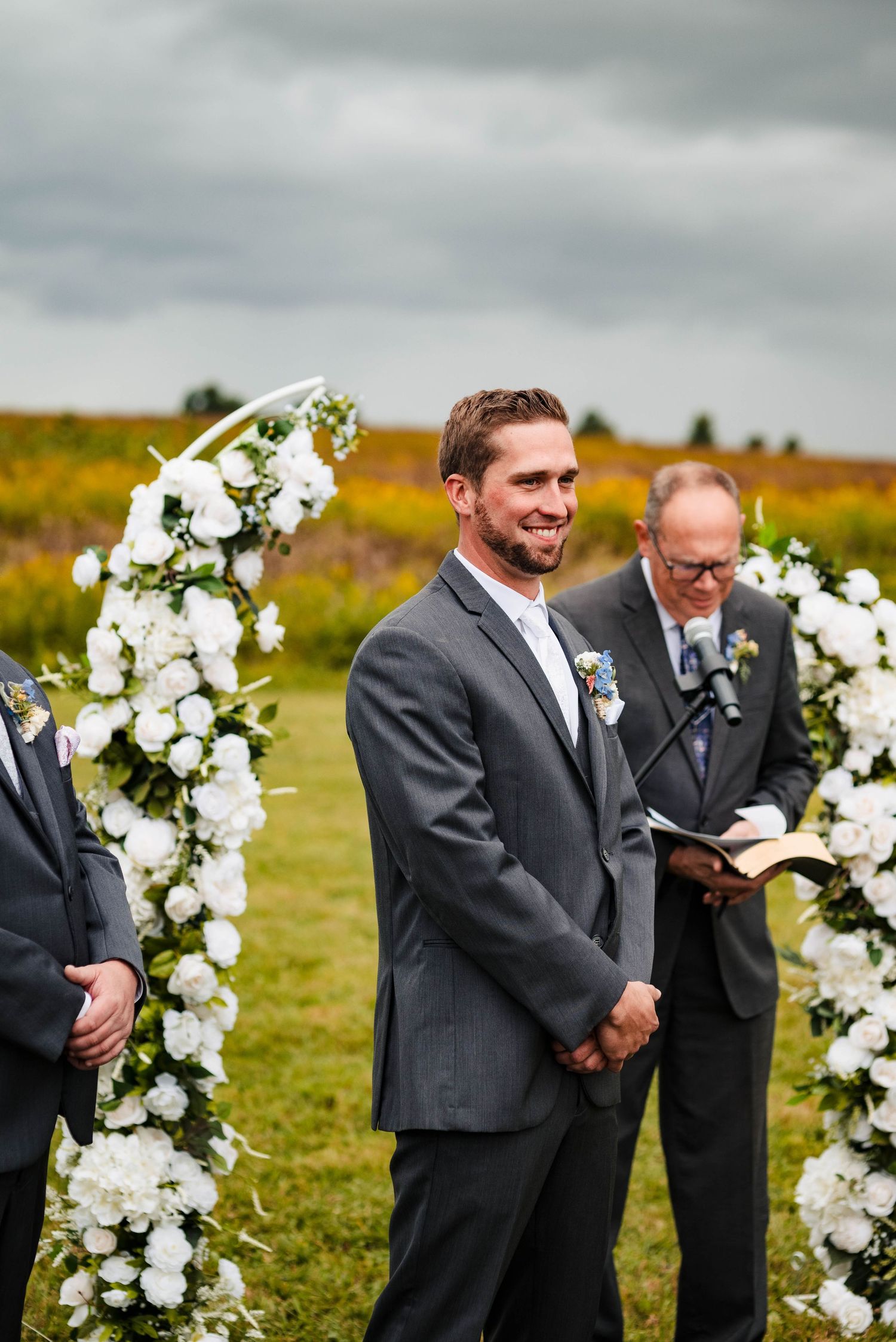 Wedding ceremony setup outdoors with white floral arch and grey skies in the background.