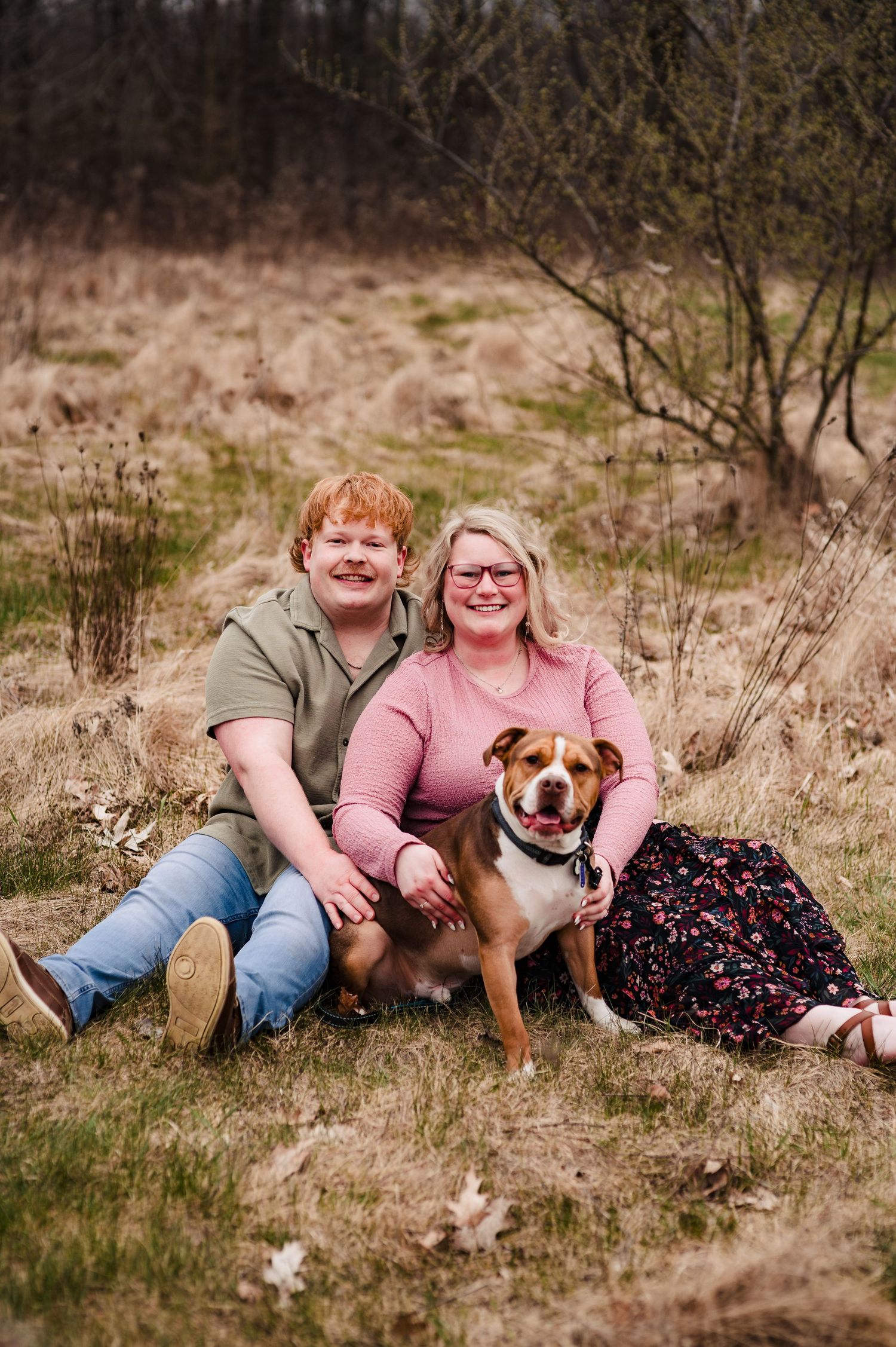 A couple sits in a field with their brown and white dog during an outdoor photo session.