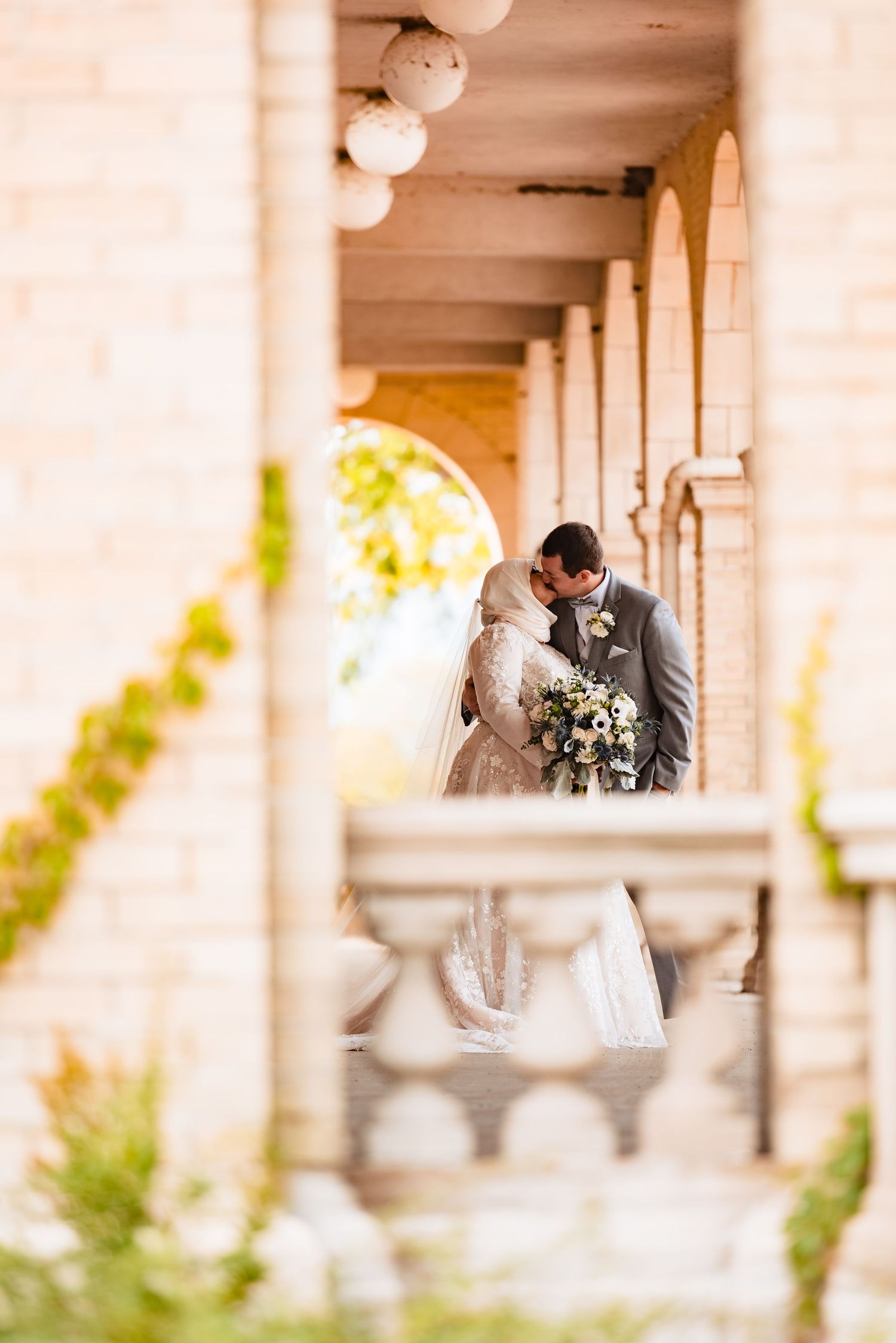 Elegant wedding couple embraces in a sunlit colonnade with white columns and romantic floral bouquet details.
