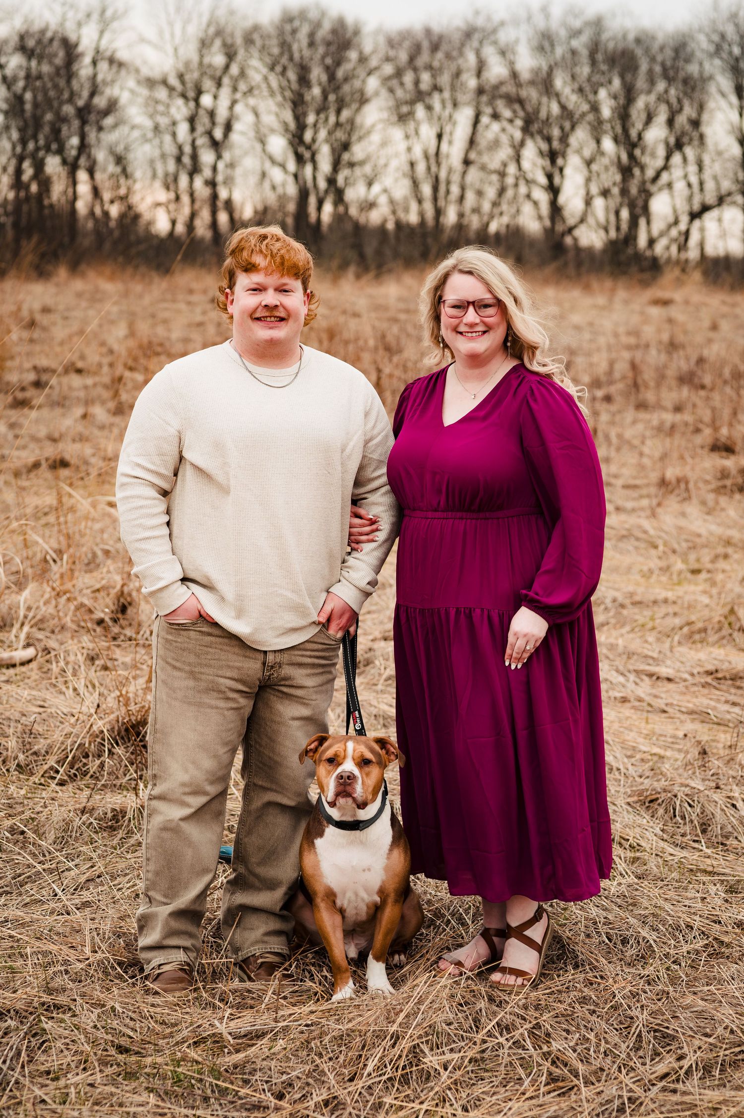 A couple stands with their dog in a winter field with leafless trees behind them.