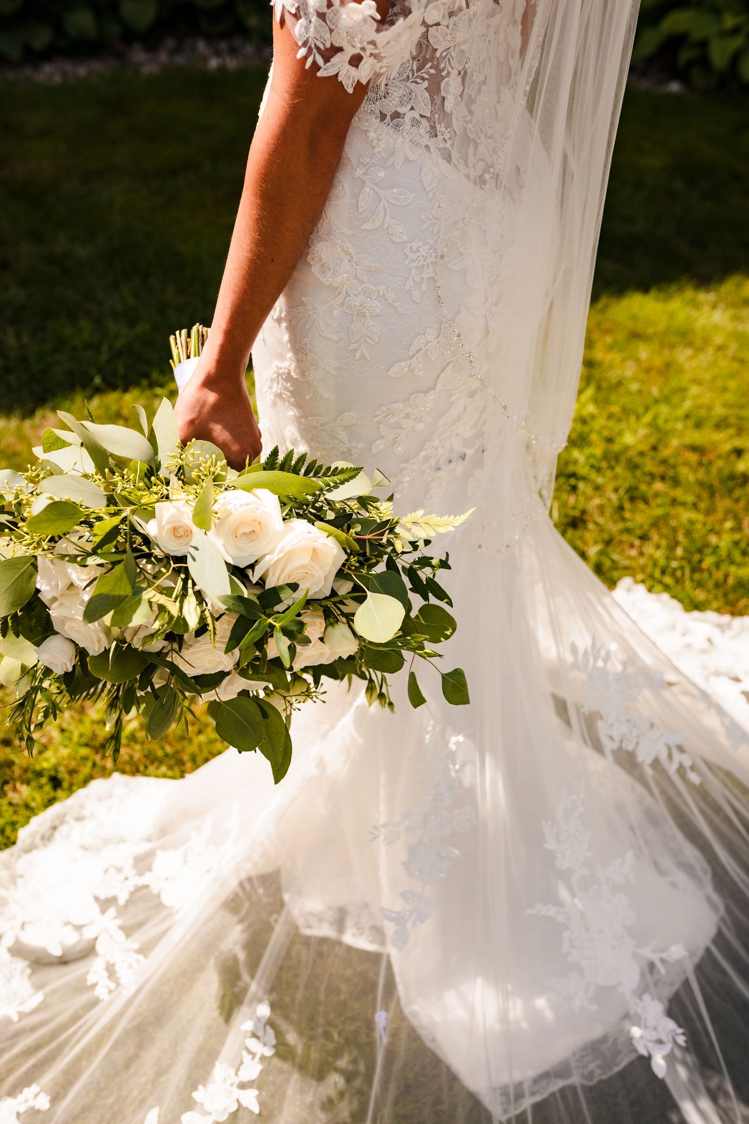 A close-up of a bride's white lace wedding dress and flowing veil with a beautiful white rose and greenery bouquet.