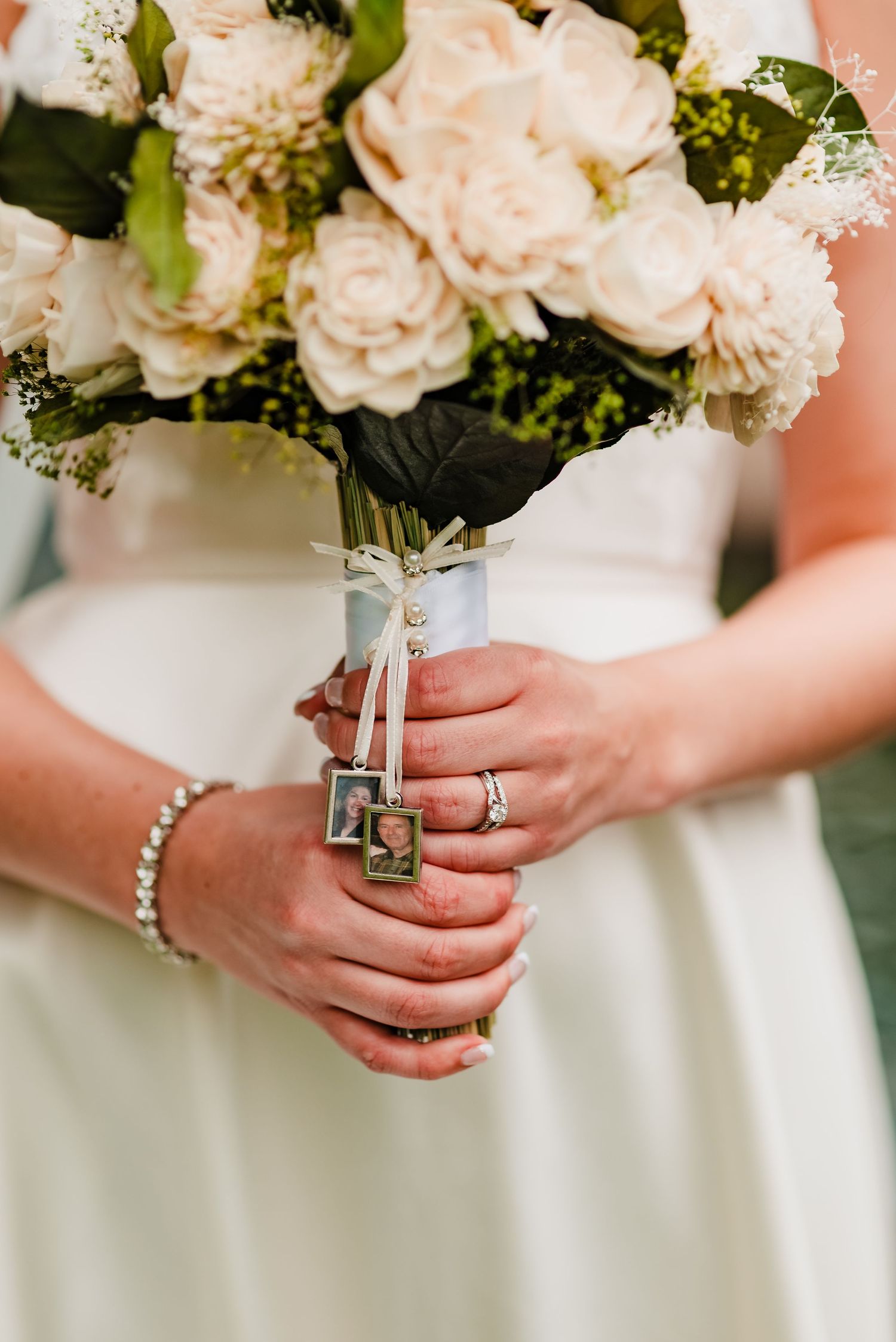 Hands holding elegant white wedding bouquet with roses and bracelets showing wedding rings and jewelry details.