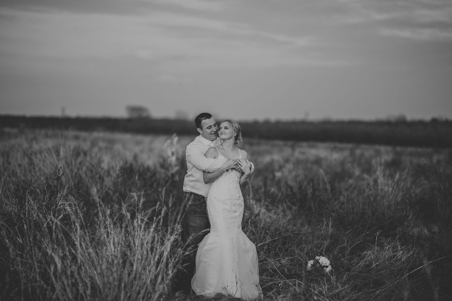 A romantic black and white wedding scene with a couple embracing in a field of tall grass and wildflowers.