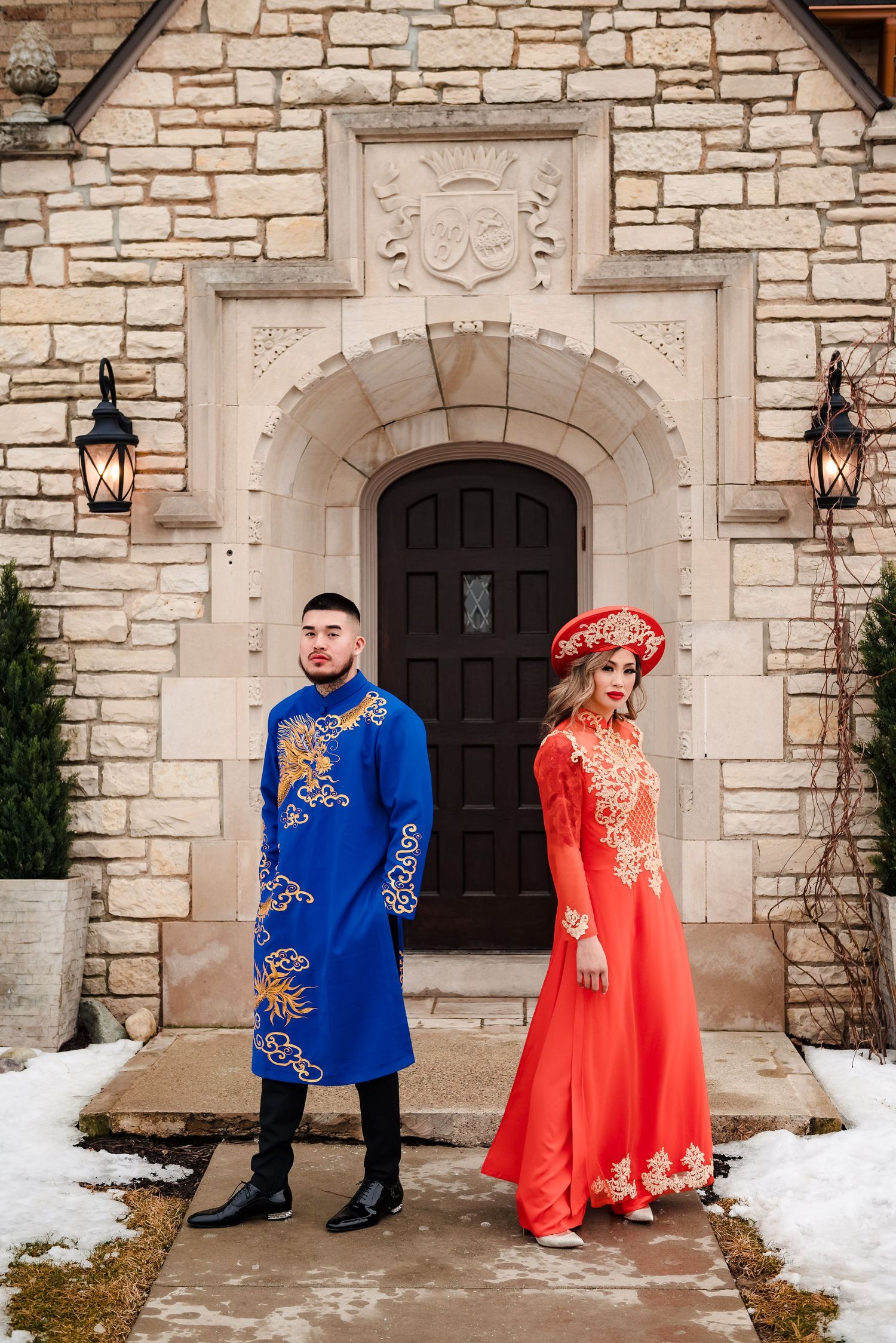Couple in vibrant blue and red traditional attire pose together against stone wall backdrop with snow on ground.