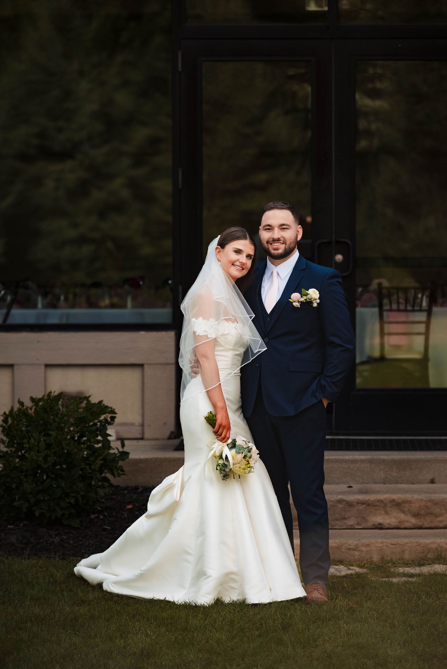 Wedding couple poses on lawn outside elegant stone venue at dusk.