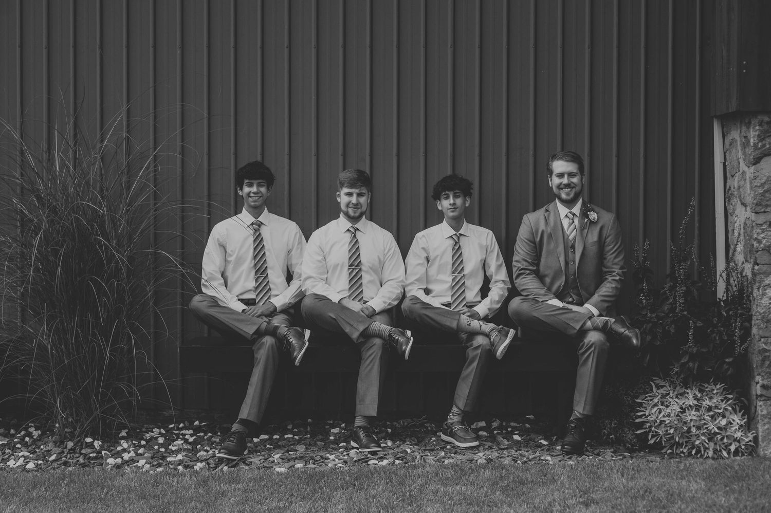 Group poses casually against dark wooden barn wall during wedding celebration.