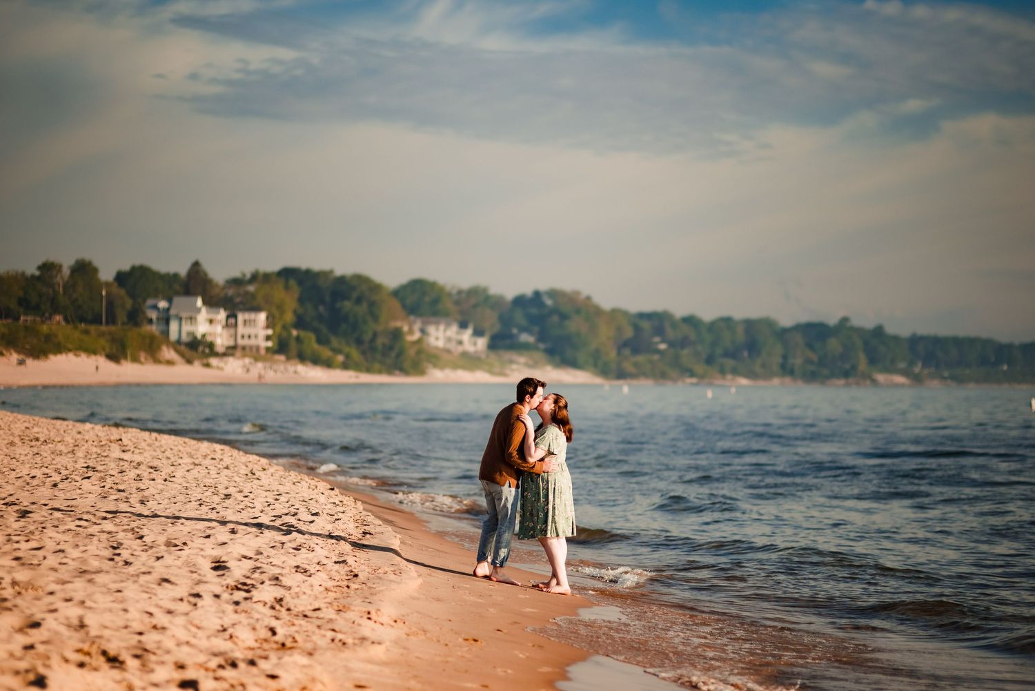 A couple embraces on a sandy beach with coastal homes visible in the background.
