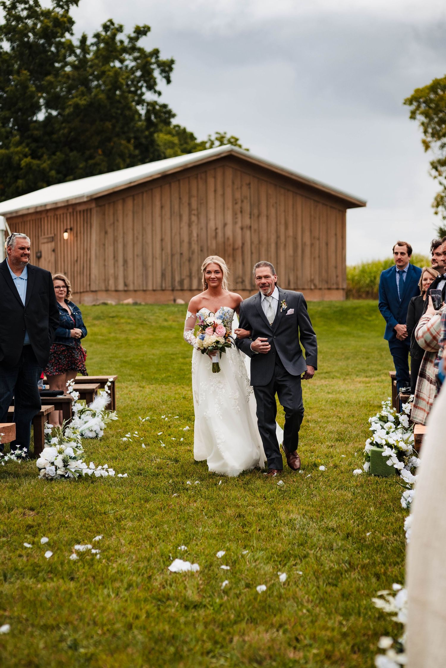 Outdoor barn wedding ceremony with flower petals lining the aisle on grass.