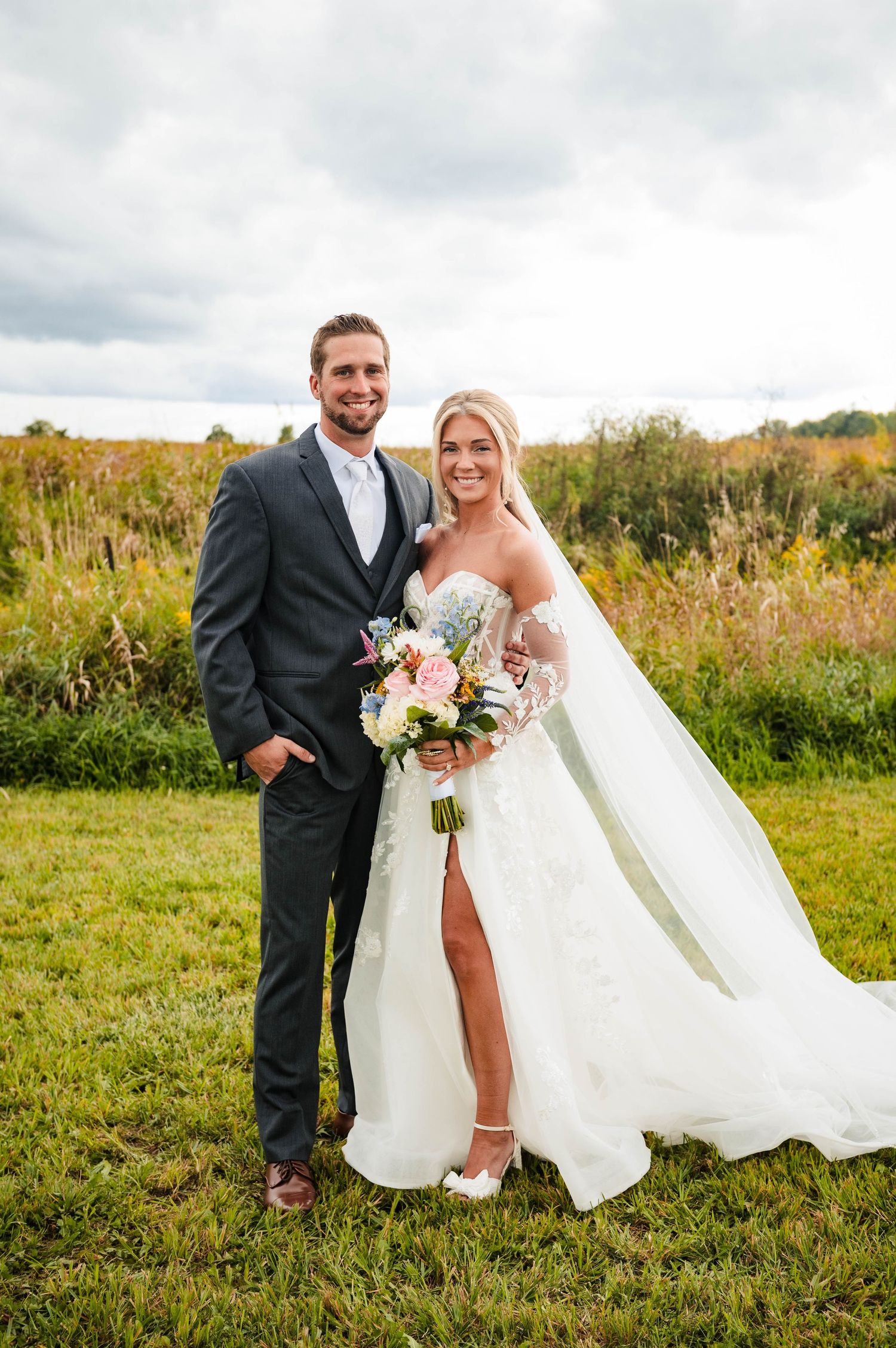 Wedding couple poses in a meadow with autumn foliage, bride holding a pink bouquet in her white gown with leg slit.