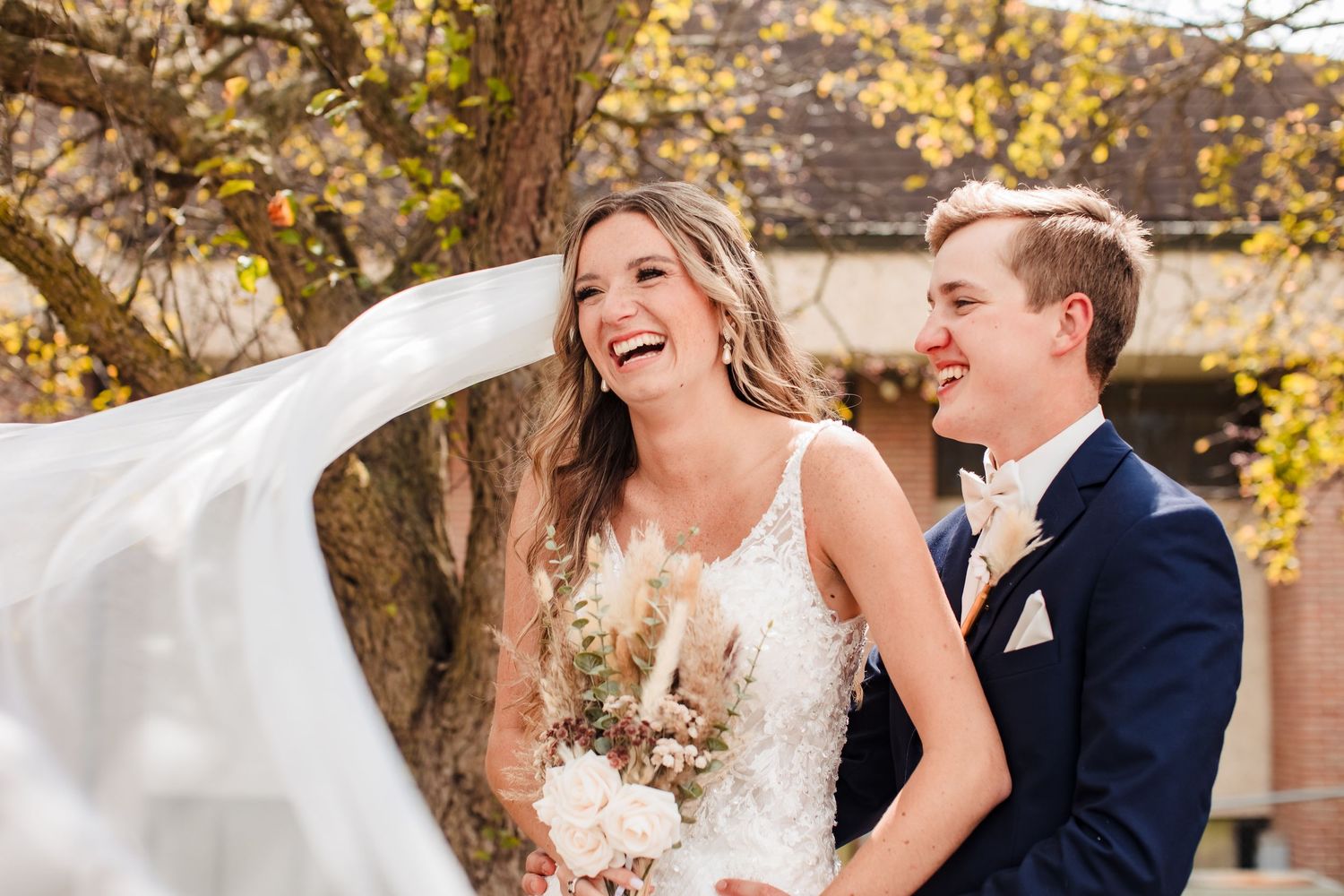 A romantic wedding couple laughs together while a veil flows in the autumn breeze during their outdoor photo session.