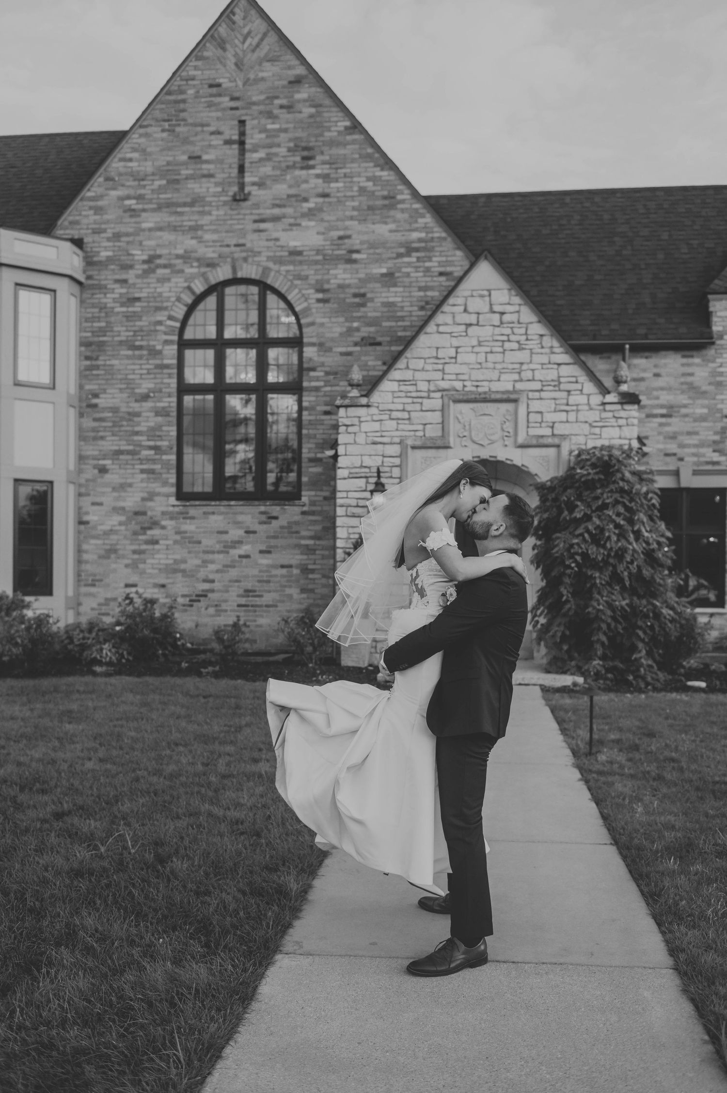 A romantic black and white portrait of newlyweds embracing outside a stone church.