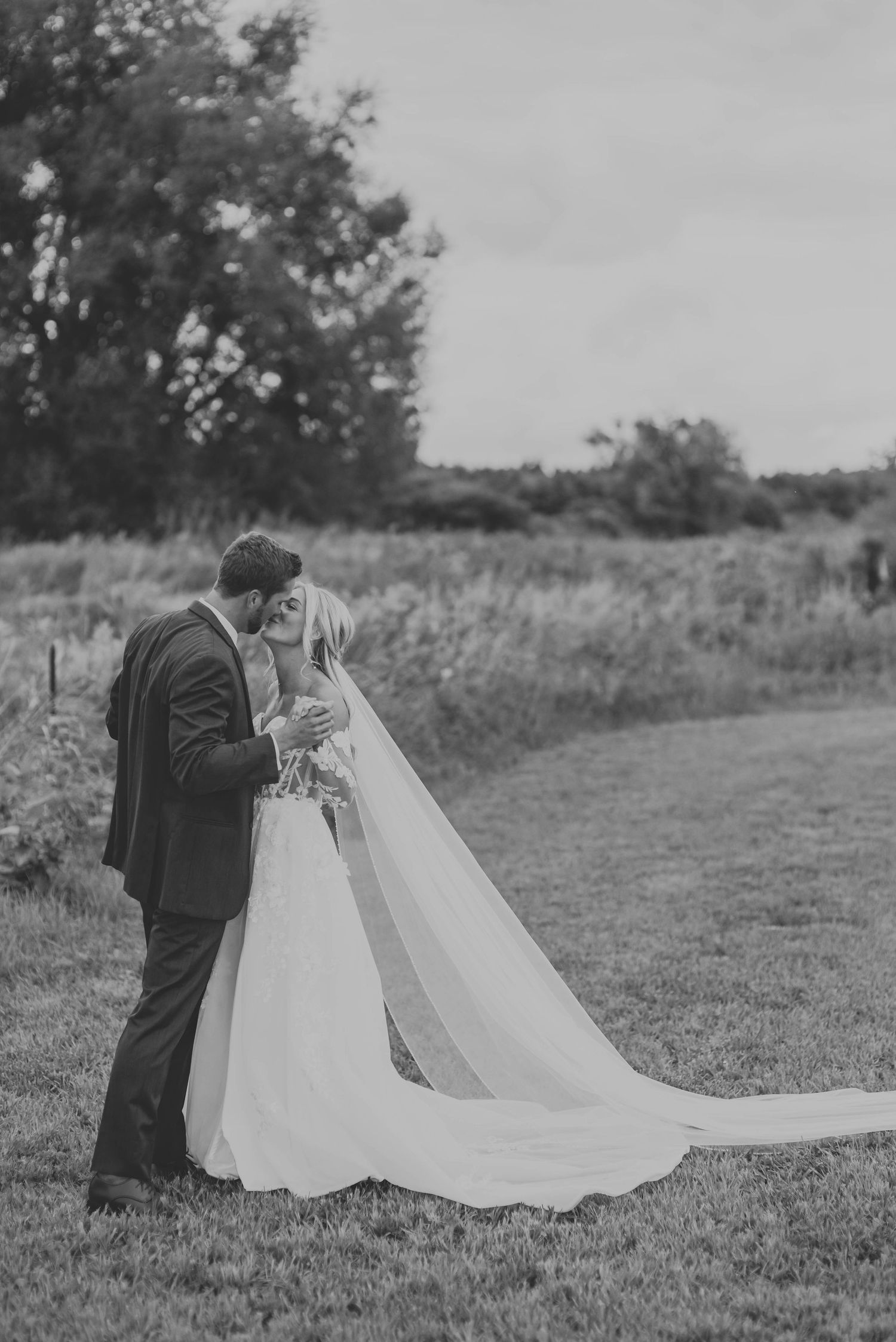 Black and white portrait of a couple in wedding attire standing close together in a meadow.