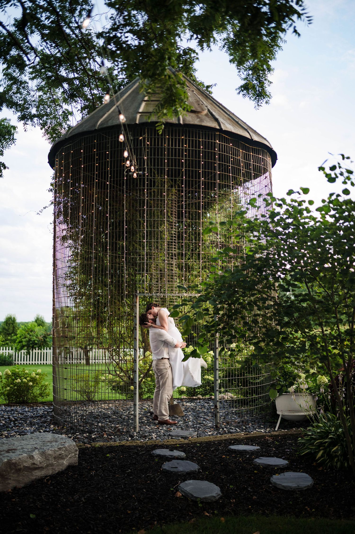 Wedding couple poses inside decorative metal gazebo surrounded by landscaped gardens.