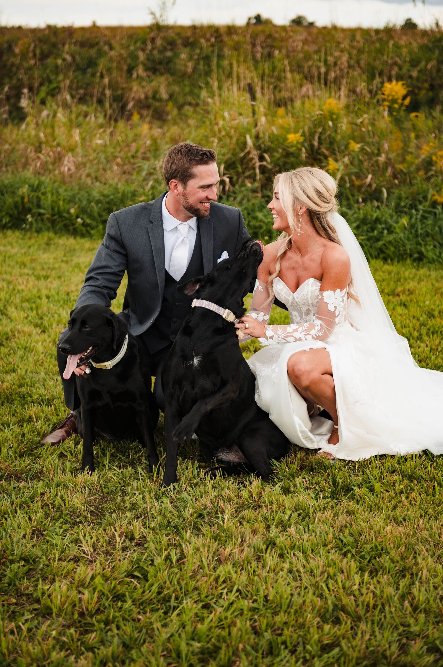 A newlywed couple sits with their black dog in a field of tall grass.