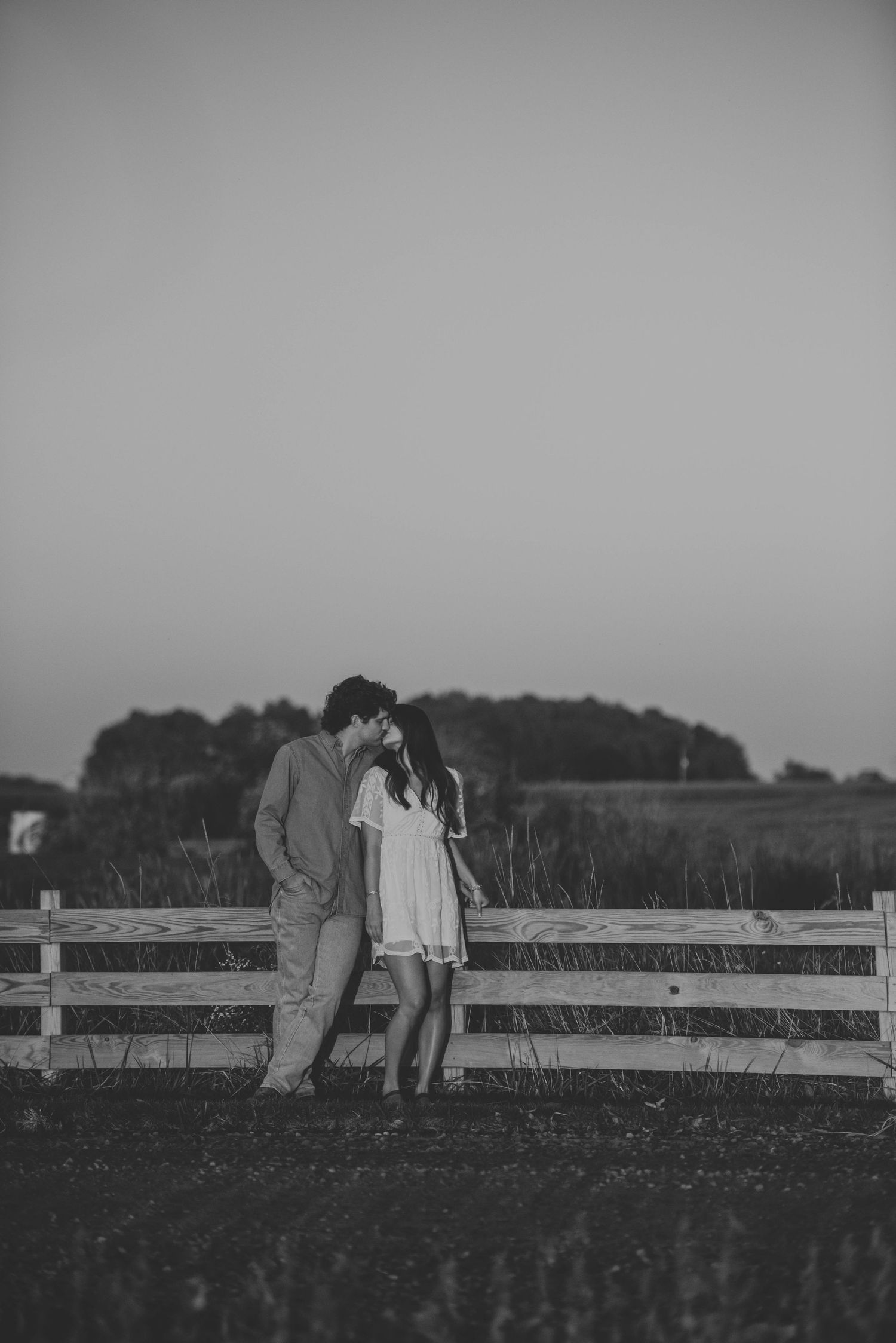 Black and white photo of a couple leaning against a wooden fence at dusk.