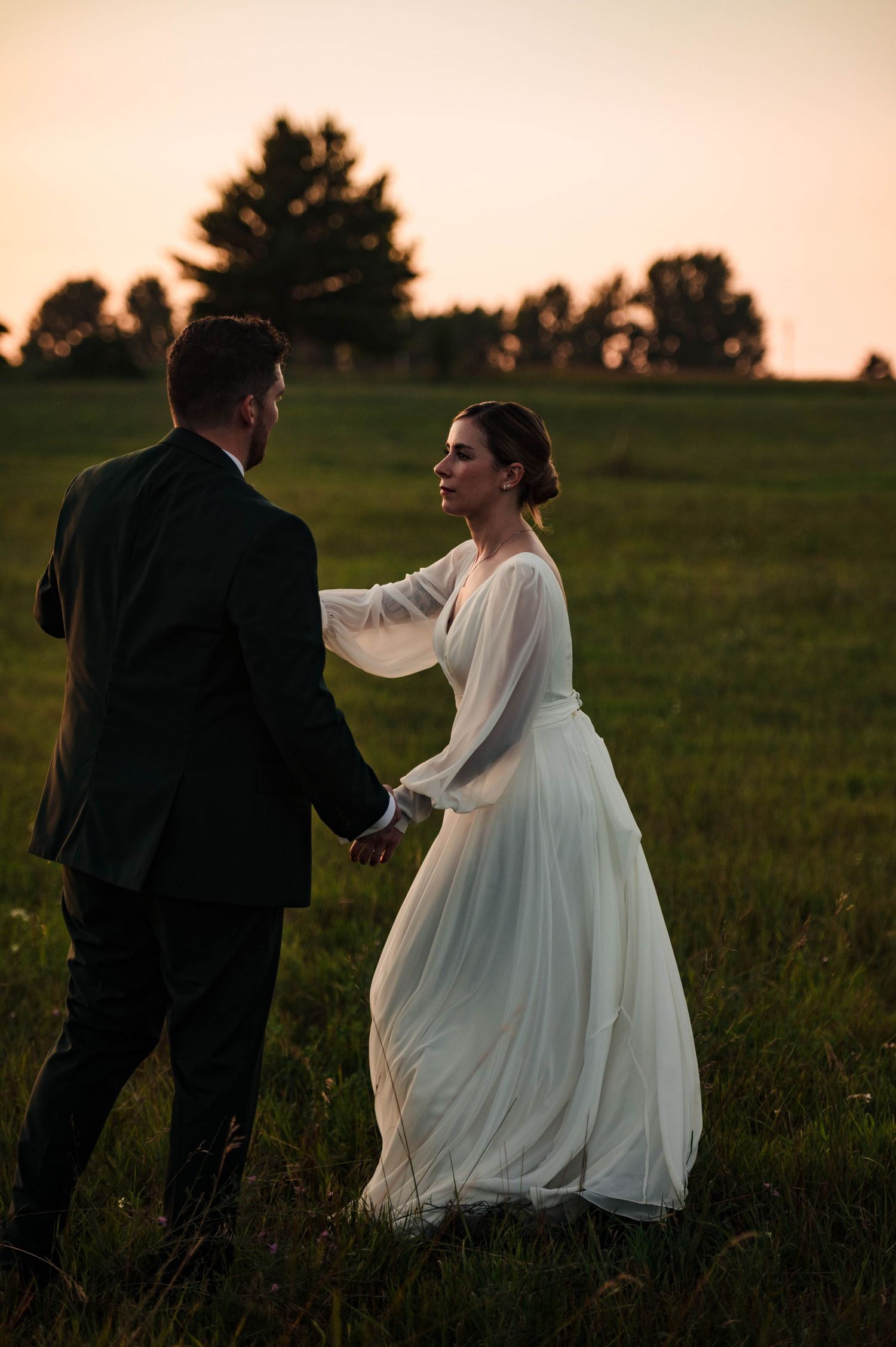 Couple in wedding attire dances in a meadow during golden hour.