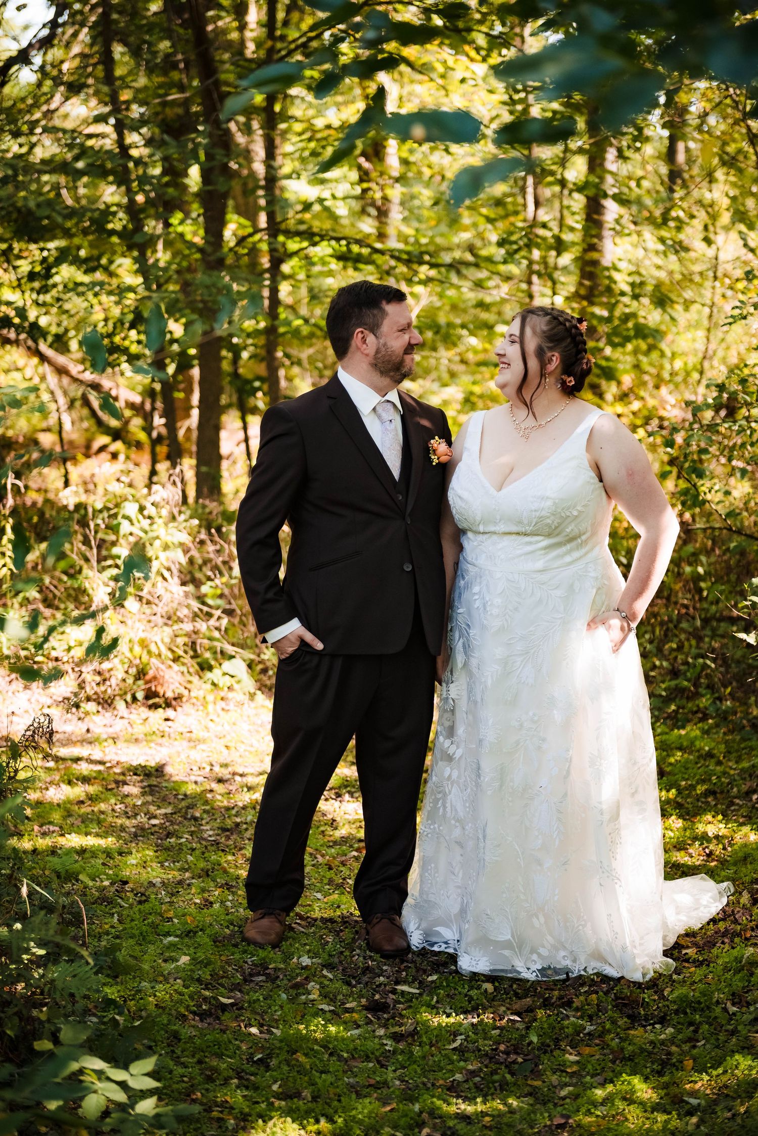 A couple poses together in formal attire against a sunlit forest backdrop.