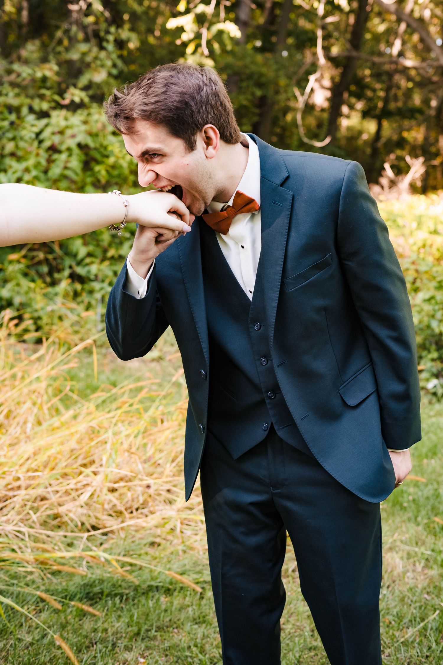 A joyful moment captured during a wedding as someone kisses their hand in a navy suit and bow tie outdoors.