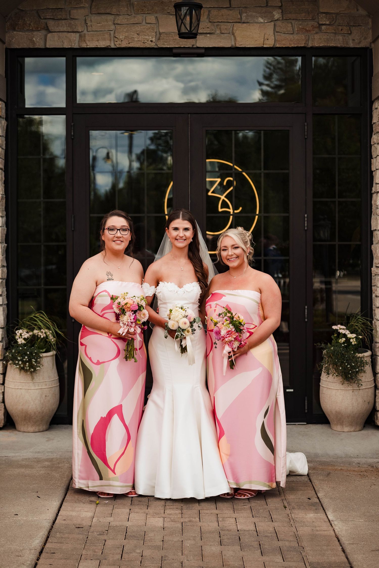 Bridesmaids in pink dresses pose with bride outside stone venue entrance.