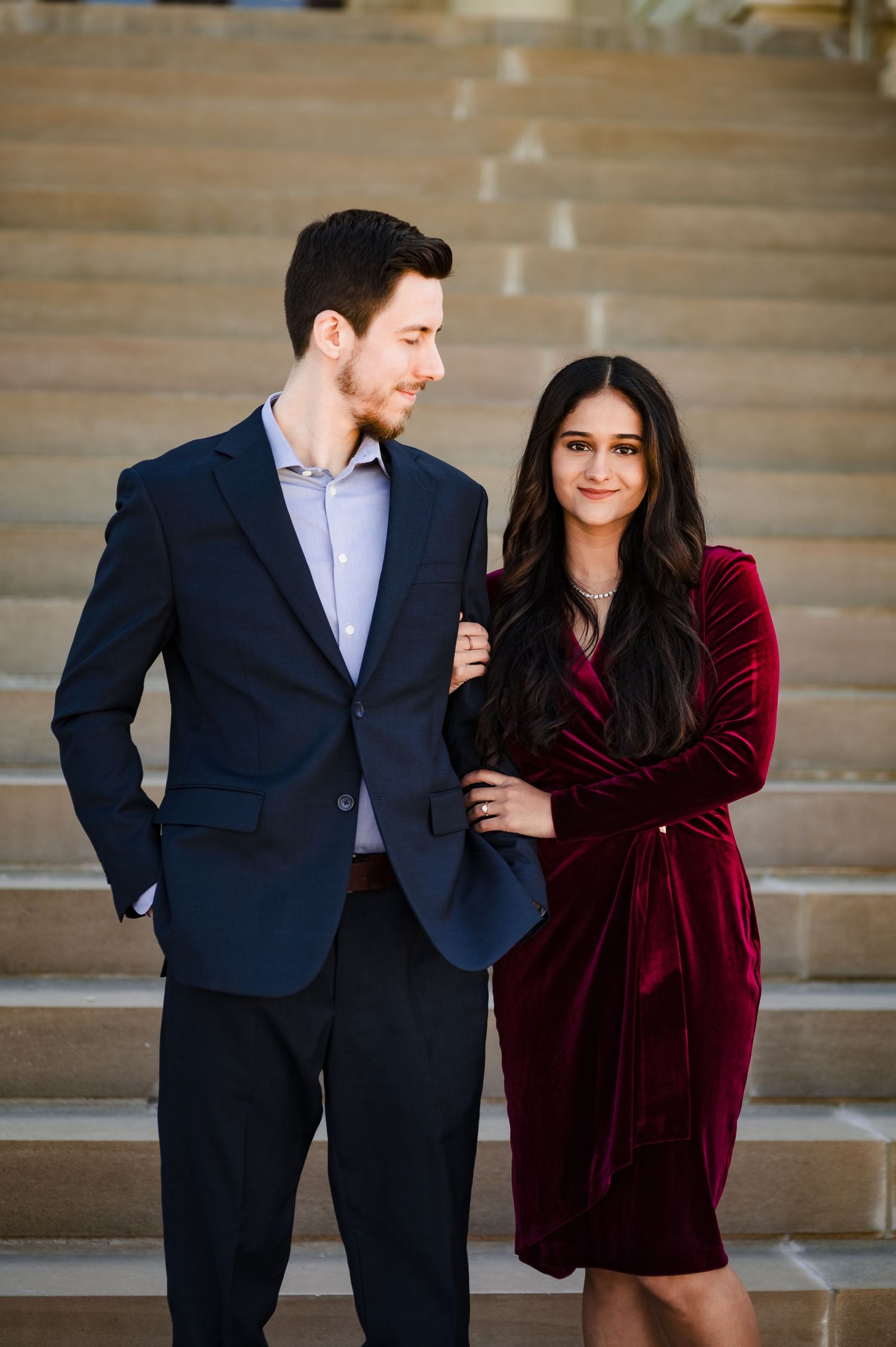 A formal couple in elegant attire poses on stone steps with a burgundy velvet dress and navy suit.