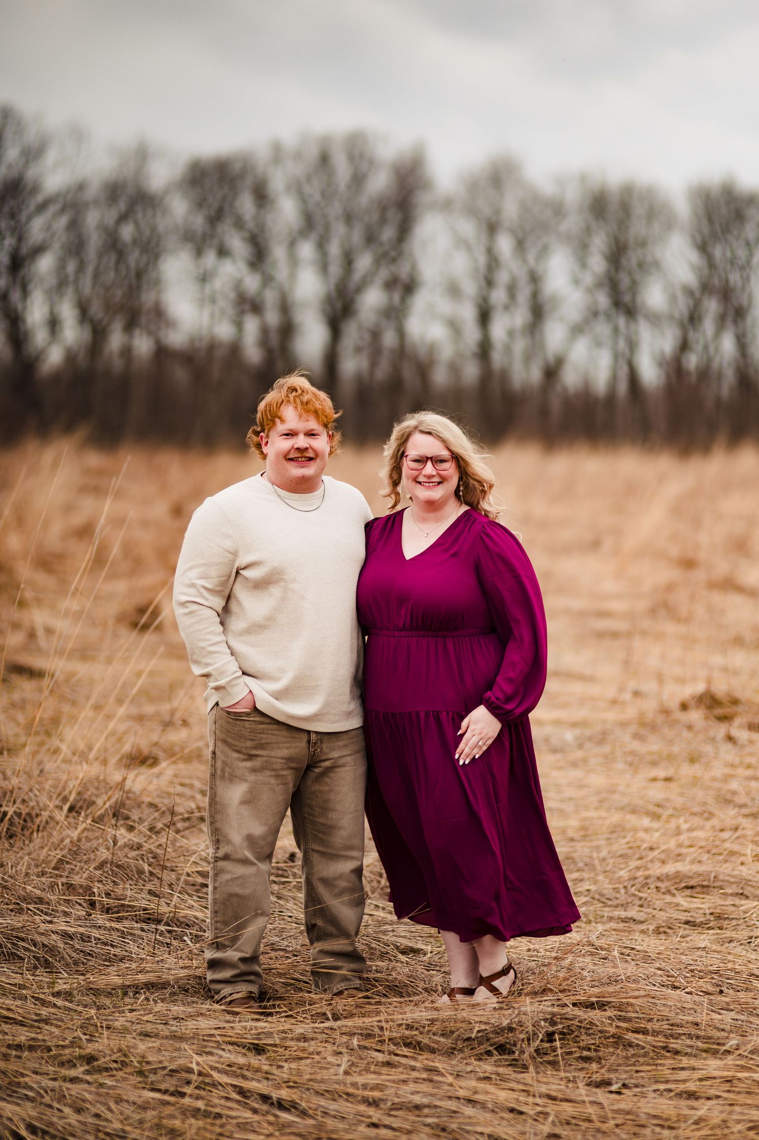 A couple poses together in a rustic field with bare trees in the background.
