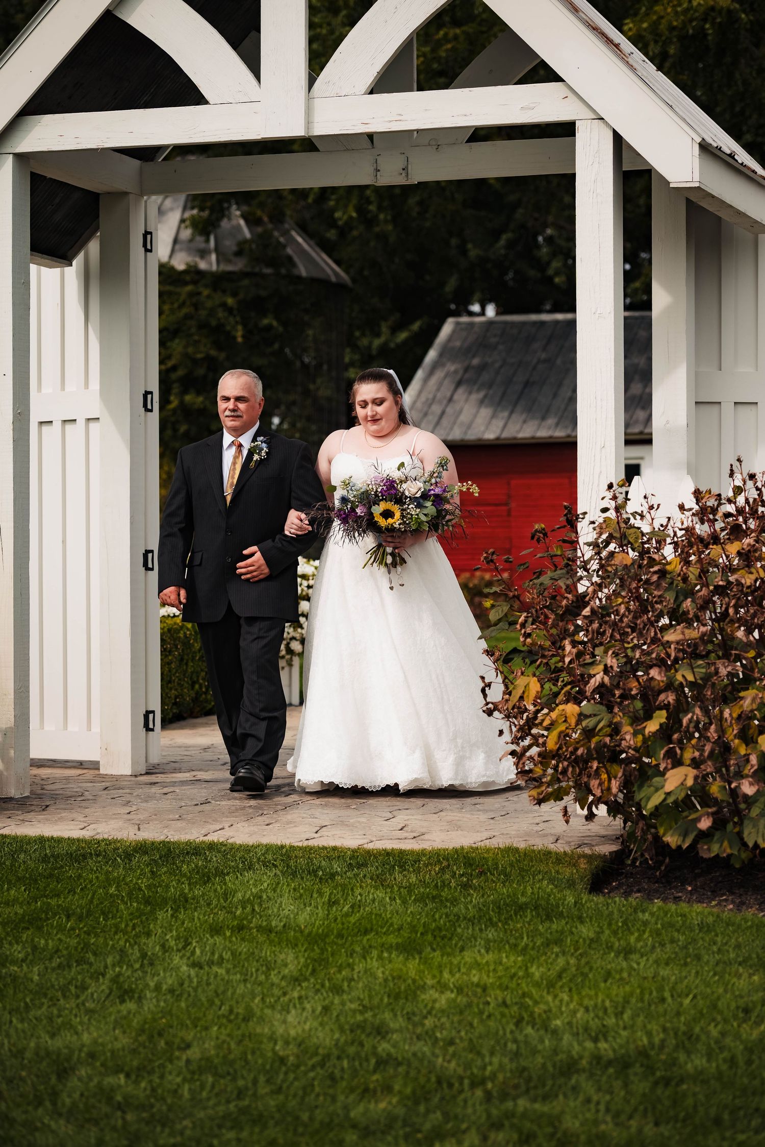 A wedding processional walks along a white pergola with a red barn backdrop.
