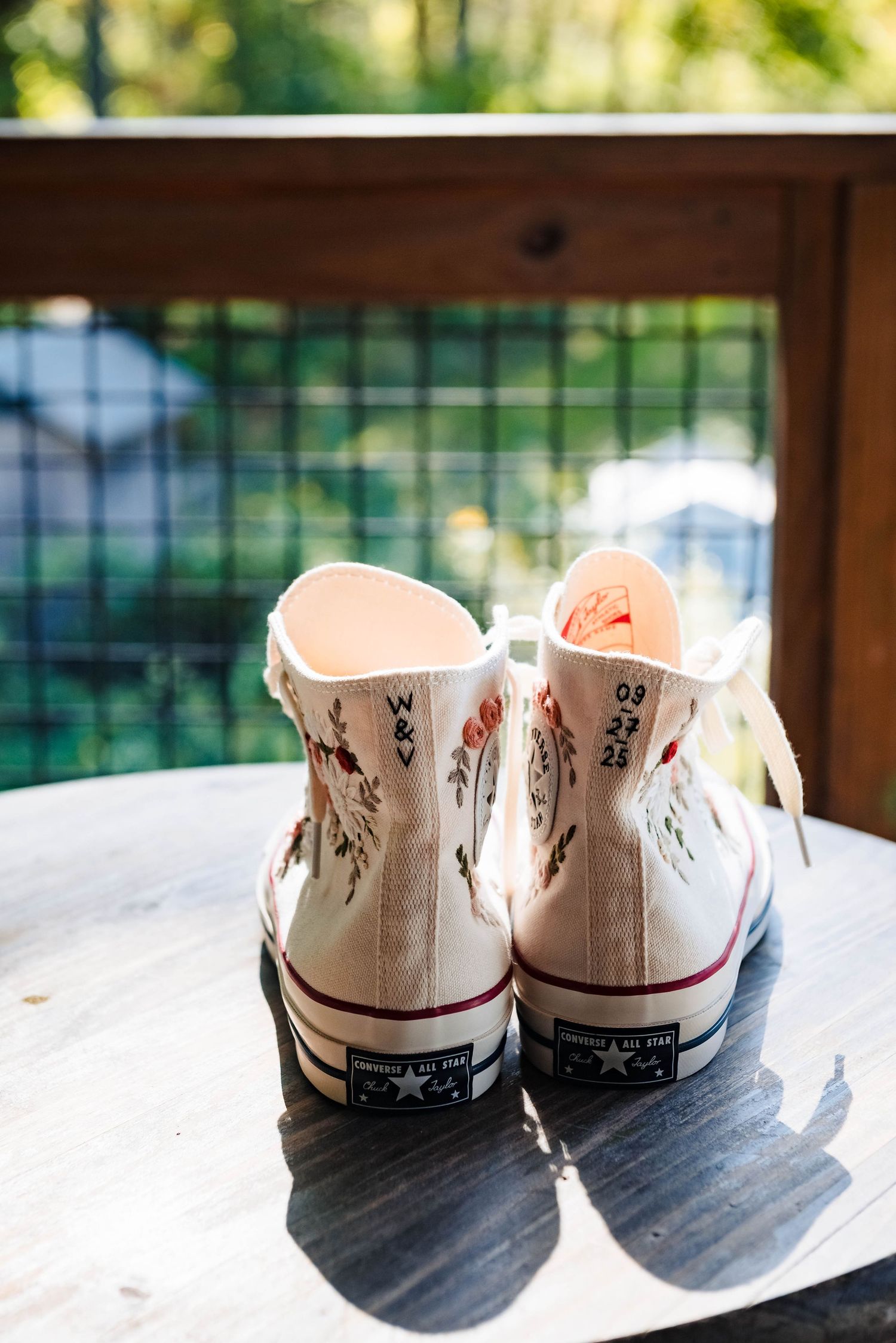White Converse sneakers decorated with floral designs casting shadows on a wooden surface.