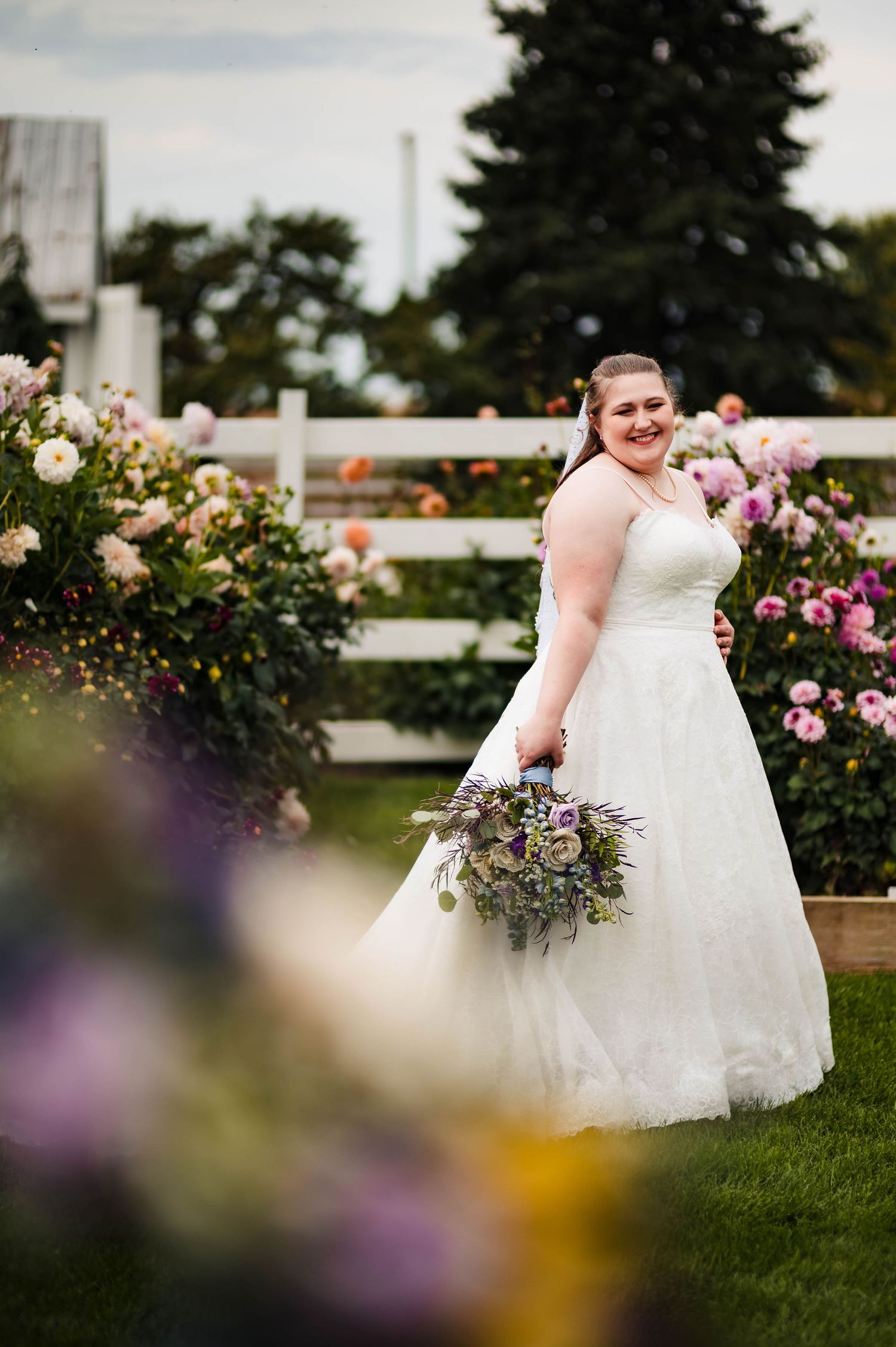 A bride in a white gown poses in a garden filled with blooming roses and colorful flowers.