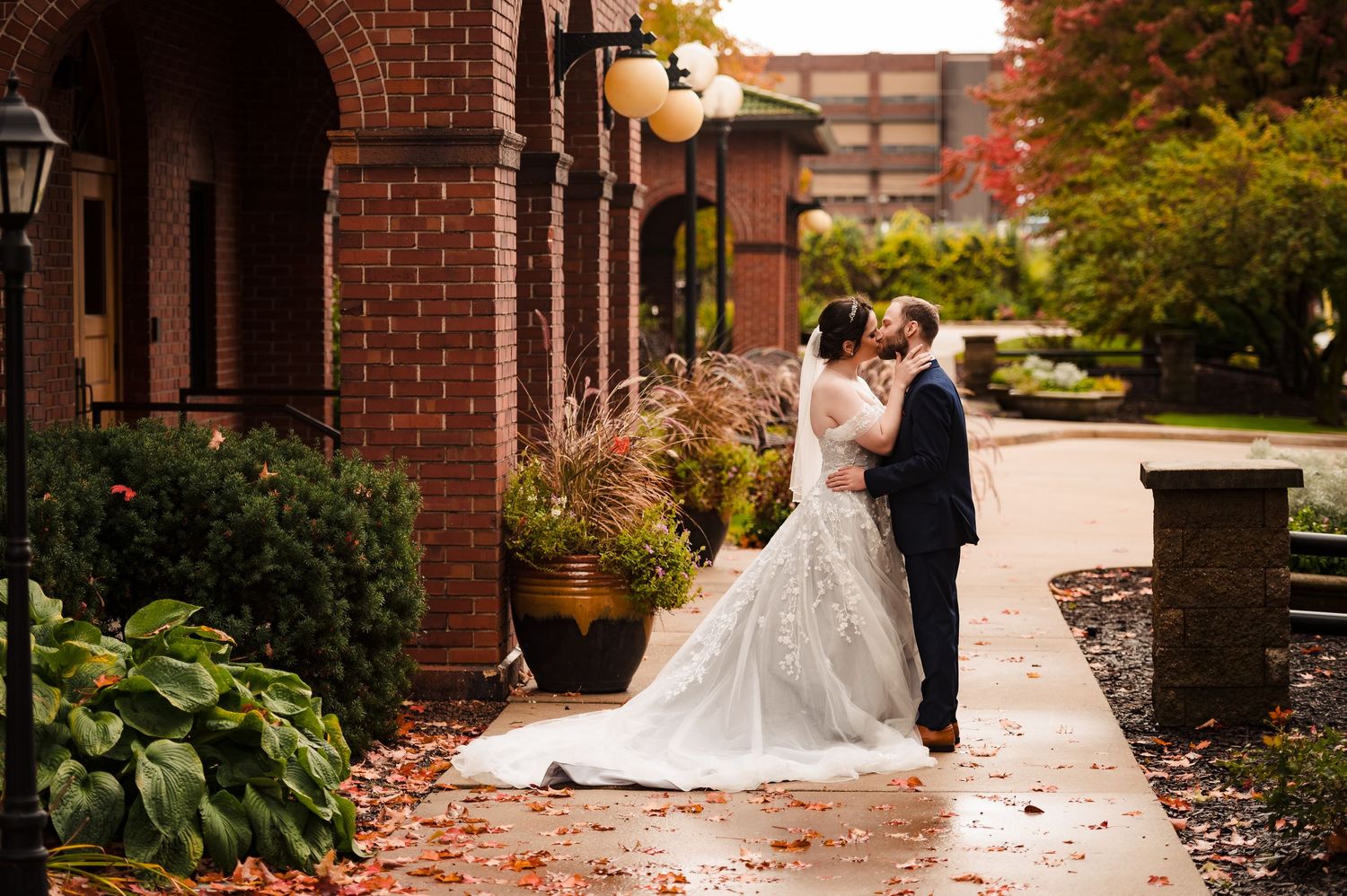 A romantic wedding couple shares a tender moment on a brick pathway surrounded by autumn foliage and architecture.