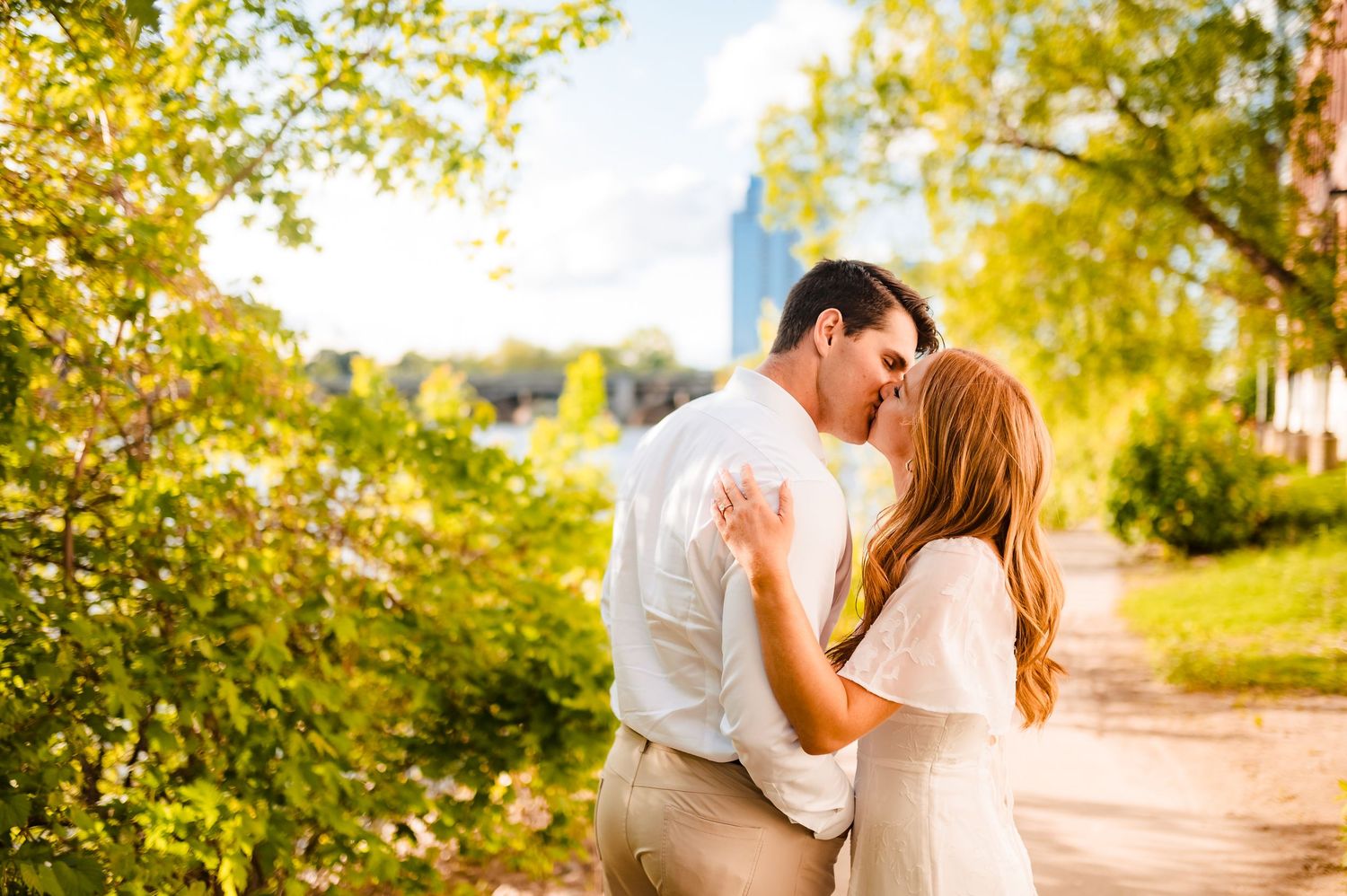 A romantic couple shares a tender kiss on a sunlit dirt path surrounded by lush summer greenery.