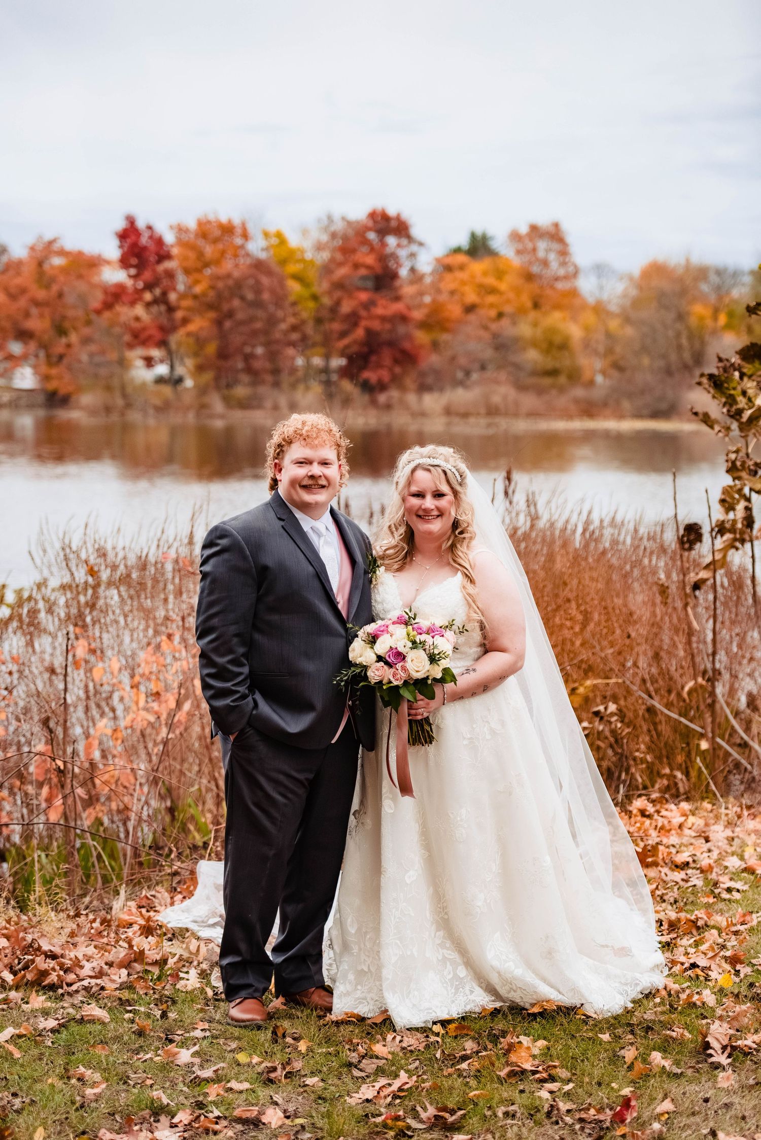 Couple laughs together during autumn wedding photoshoot by lake with colorful fall foliage in background.