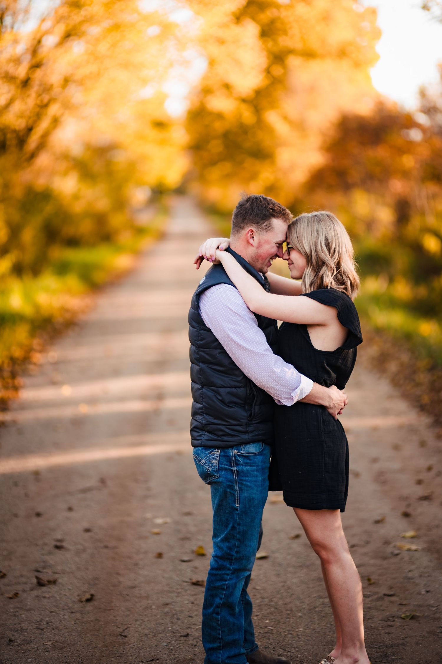 A romantic couple embraces on an autumn path surrounded by golden trees and falling leaves.