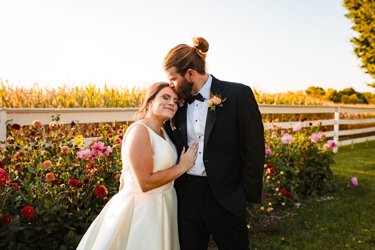 A couple in wedding attire embraces during golden hour in a garden filled with blooming roses and flowers.