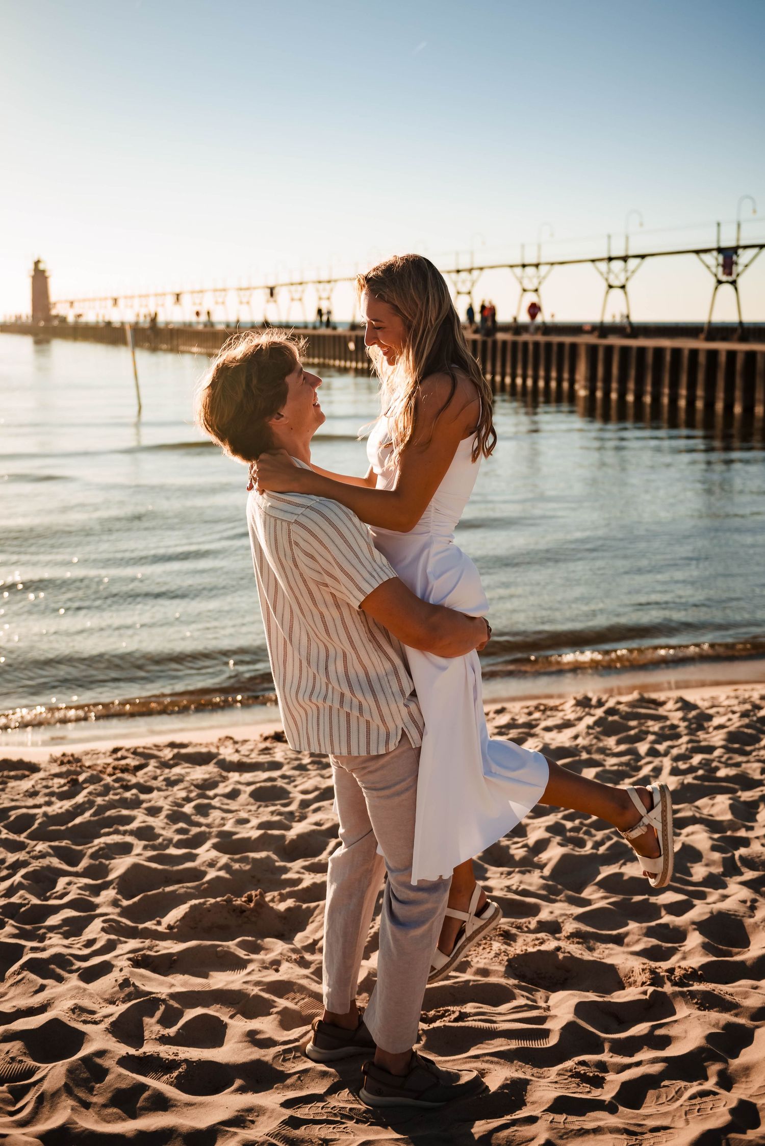 Couple embracing on sandy beach with pier and ocean view at golden hour.