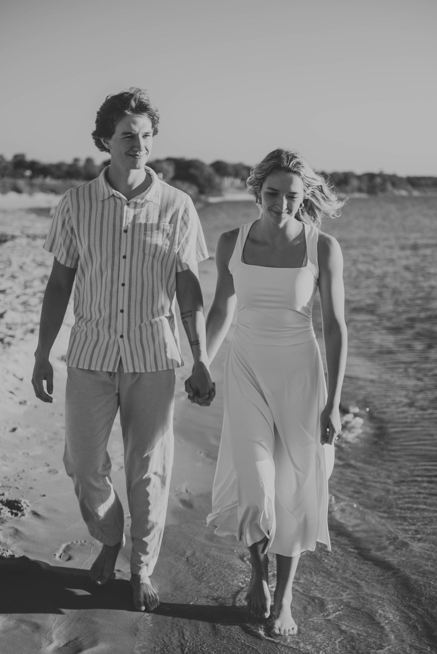 Black and white photo of couple walking along beach shoreline in summer attire.