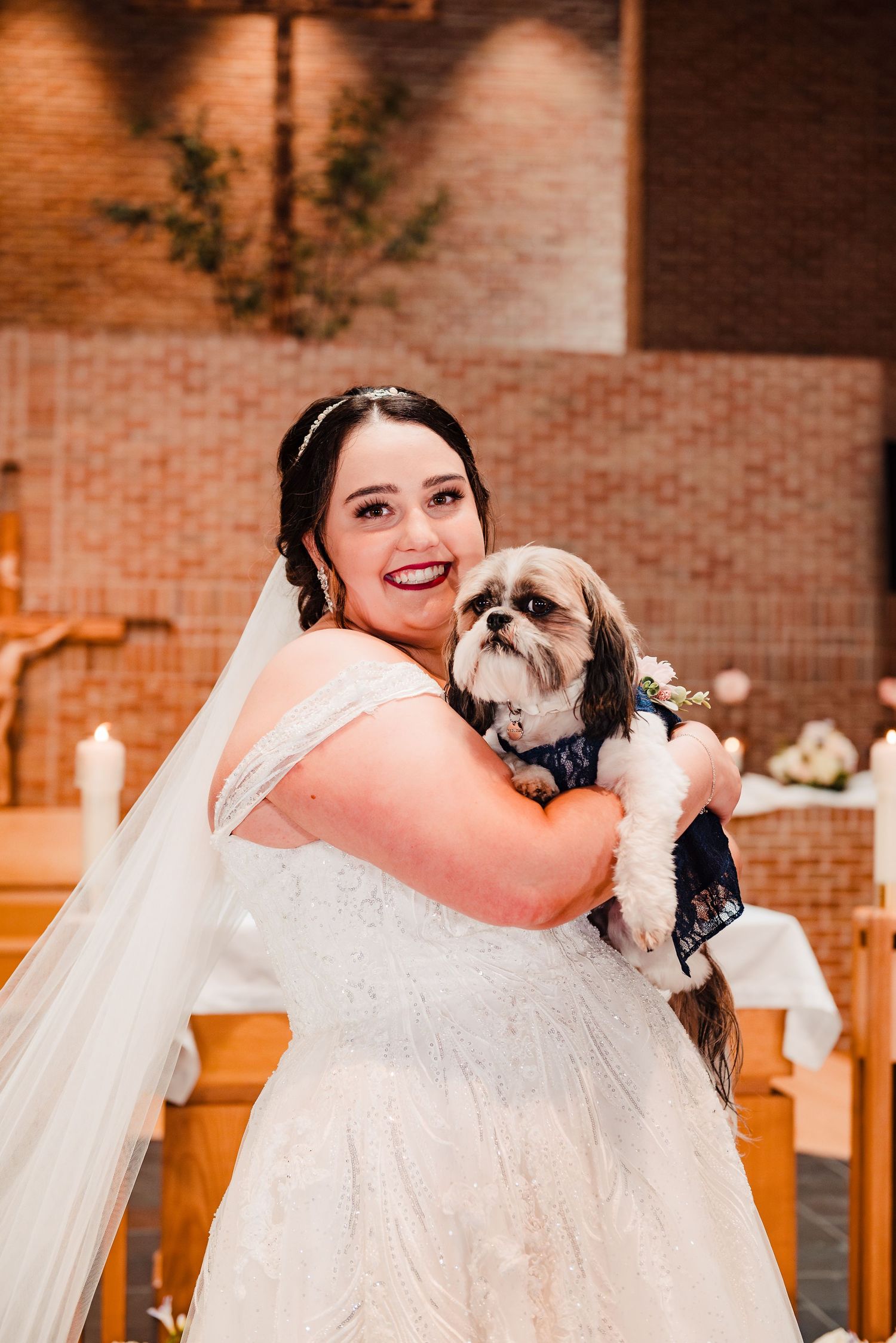 A bride in a white wedding dress shares a joyful moment with her small Shih Tzu dog during the ceremony.