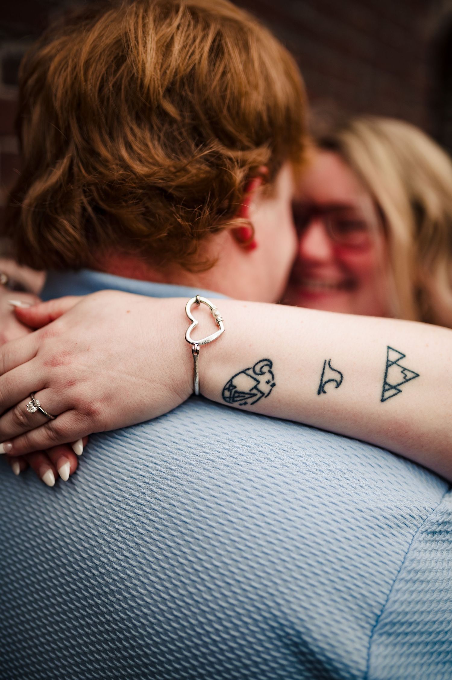 Close-up of geometric tattoos and a heart bracelet on someone's arm during an embrace.