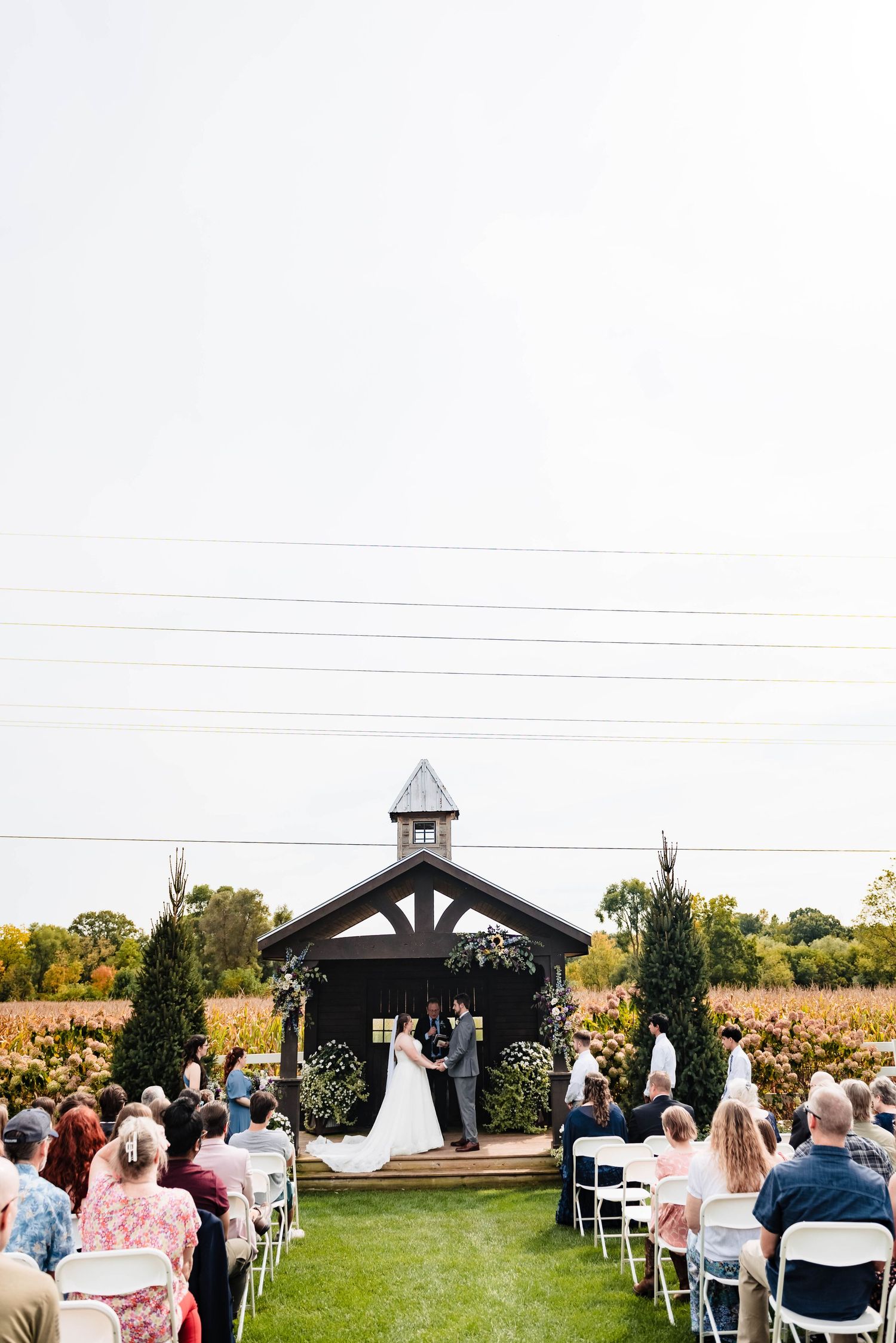 An outdoor wedding ceremony at a charming black gazebo with white chairs and guests in attendance.