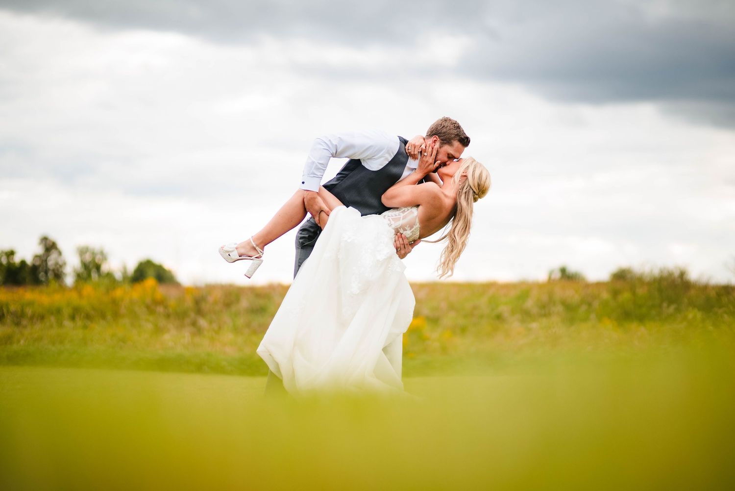 A playful wedding moment captured in a green field with dramatic cloudy sky.