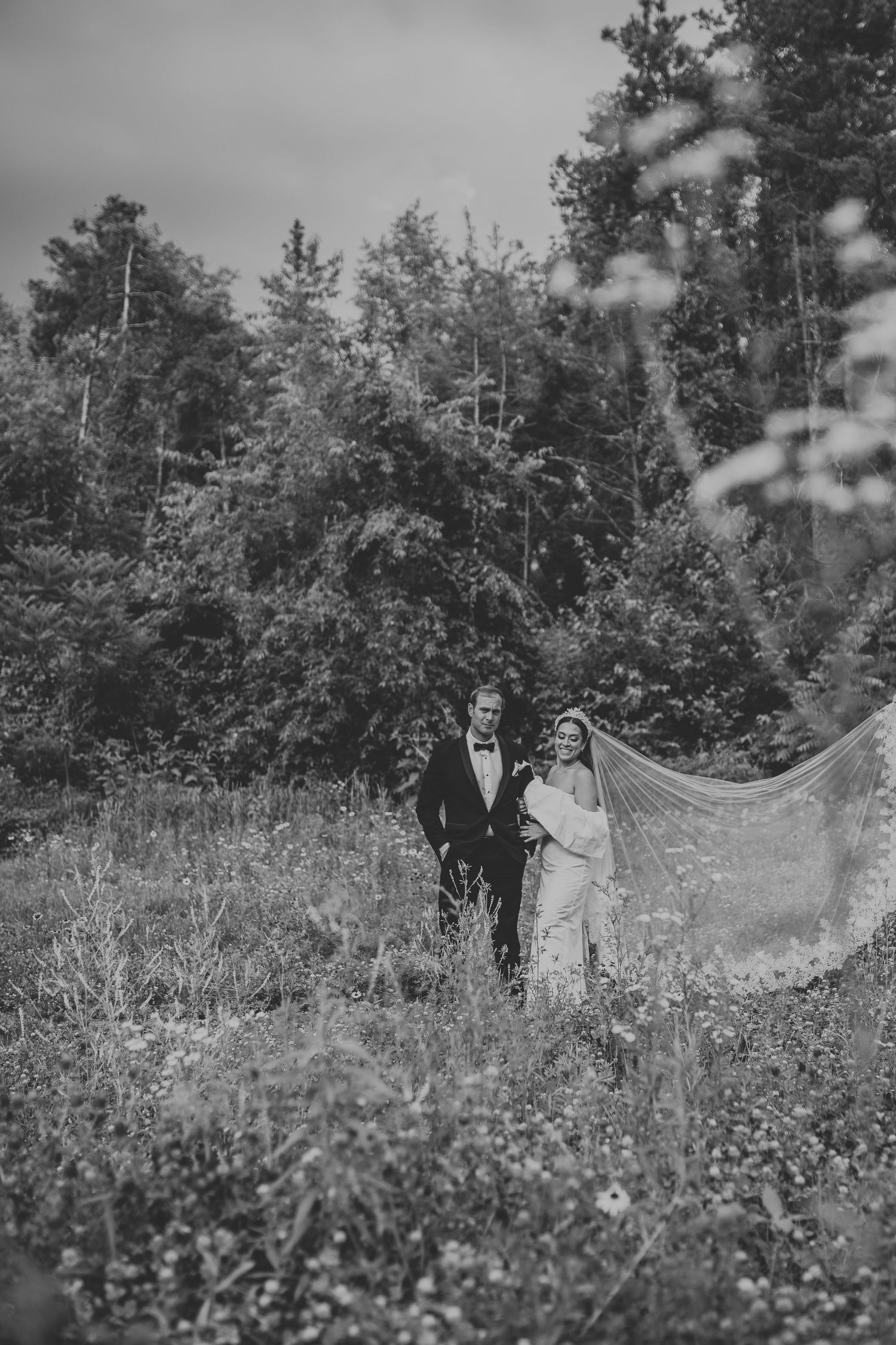 A black and white wedding photo showing a couple walking through a wildflower meadow with trees in the background.