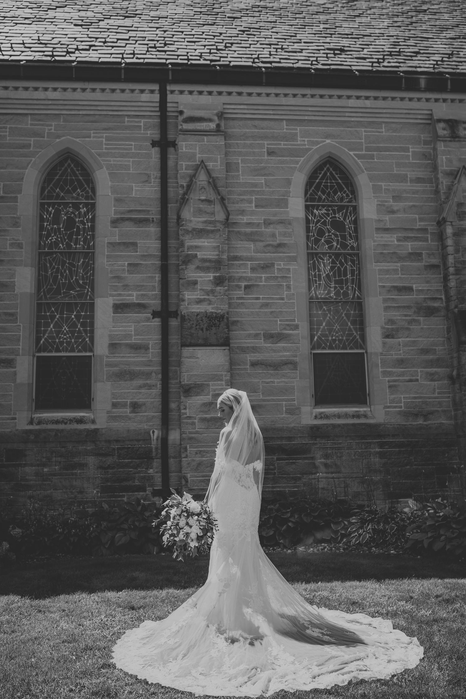 A black and white photo of a bride in a flowing lace gown and veil standing beside a gothic stone church.