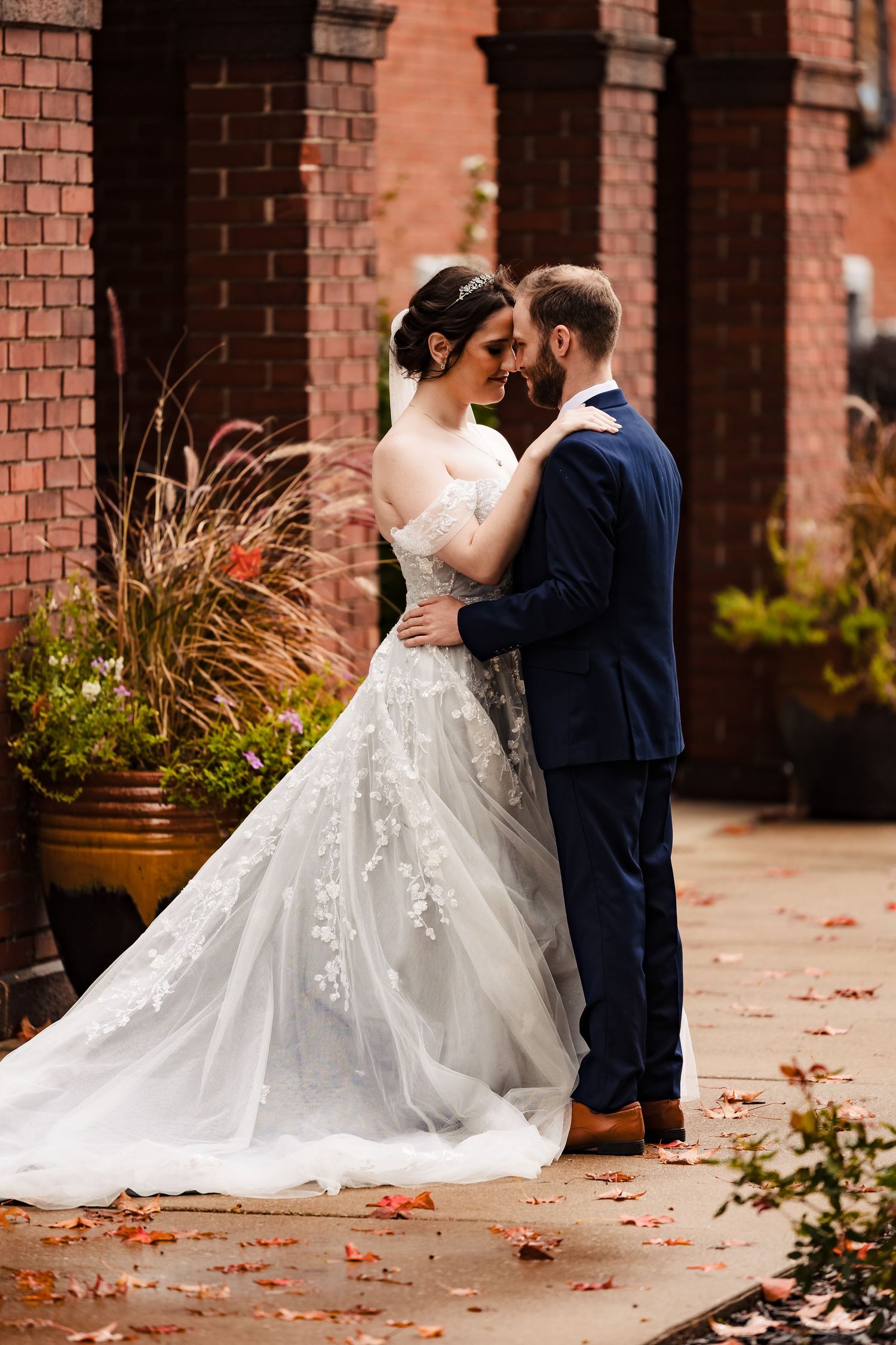 A romantic wedding couple embraces against a rustic brick wall backdrop with autumn leaves scattered on the ground.