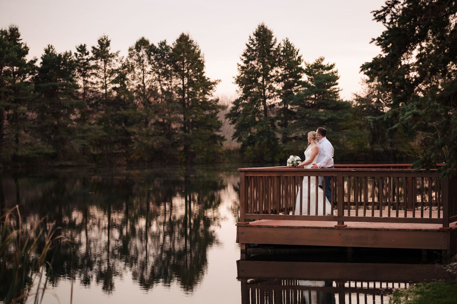 A couple shares a romantic moment on a wooden dock overlooking a serene lake at sunset with pine trees in the background.
