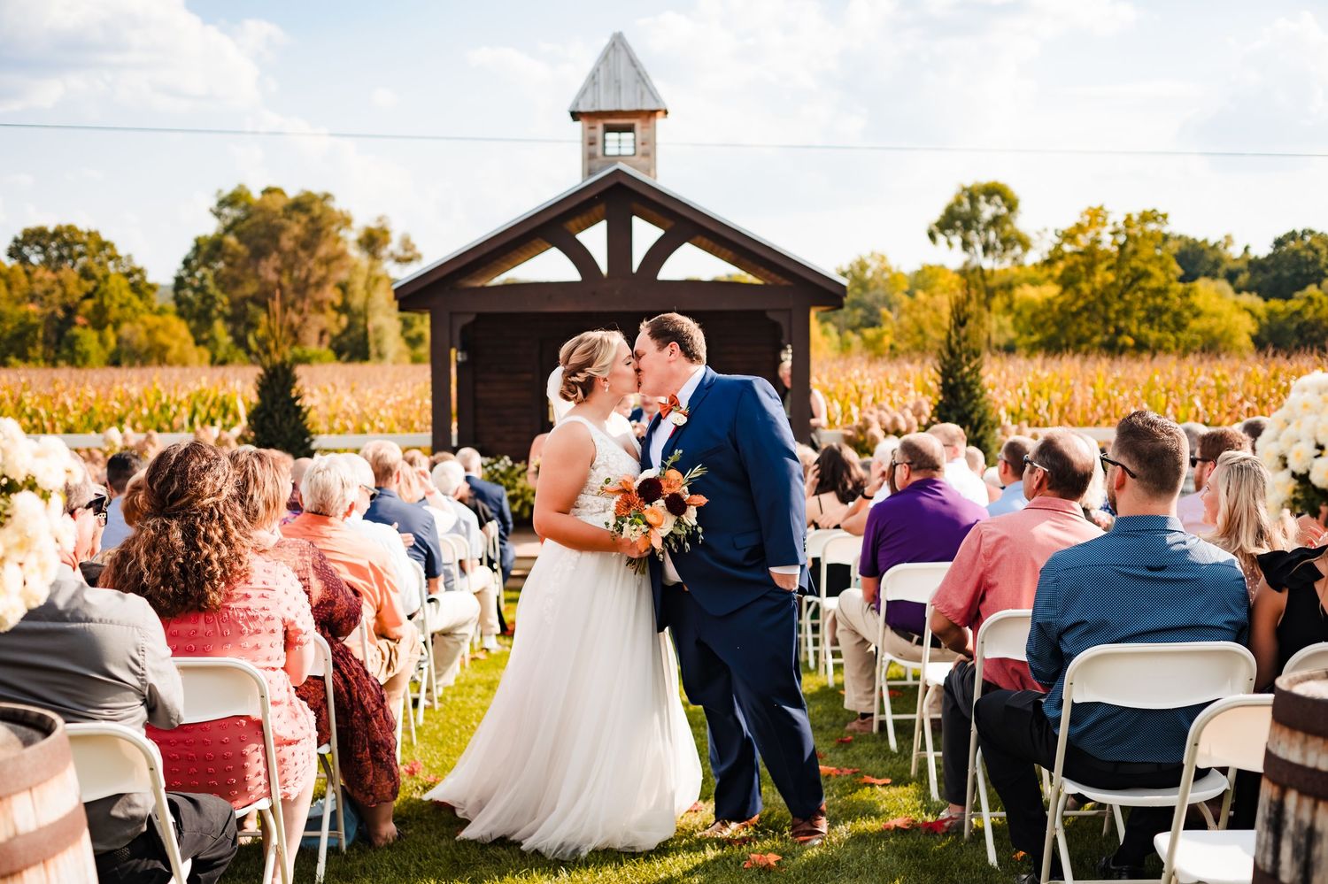 Panoramic view of an outdoor wedding ceremony at a rustic chapel surrounded by autumn trees and seated guests.