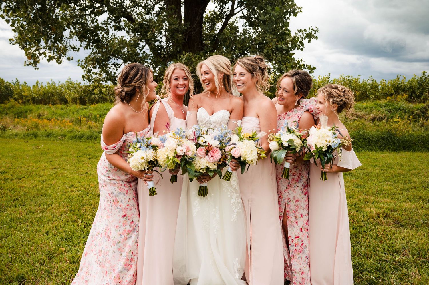 Wedding party in pink and white dresses pose with floral bouquets in a sunny outdoor field setting.