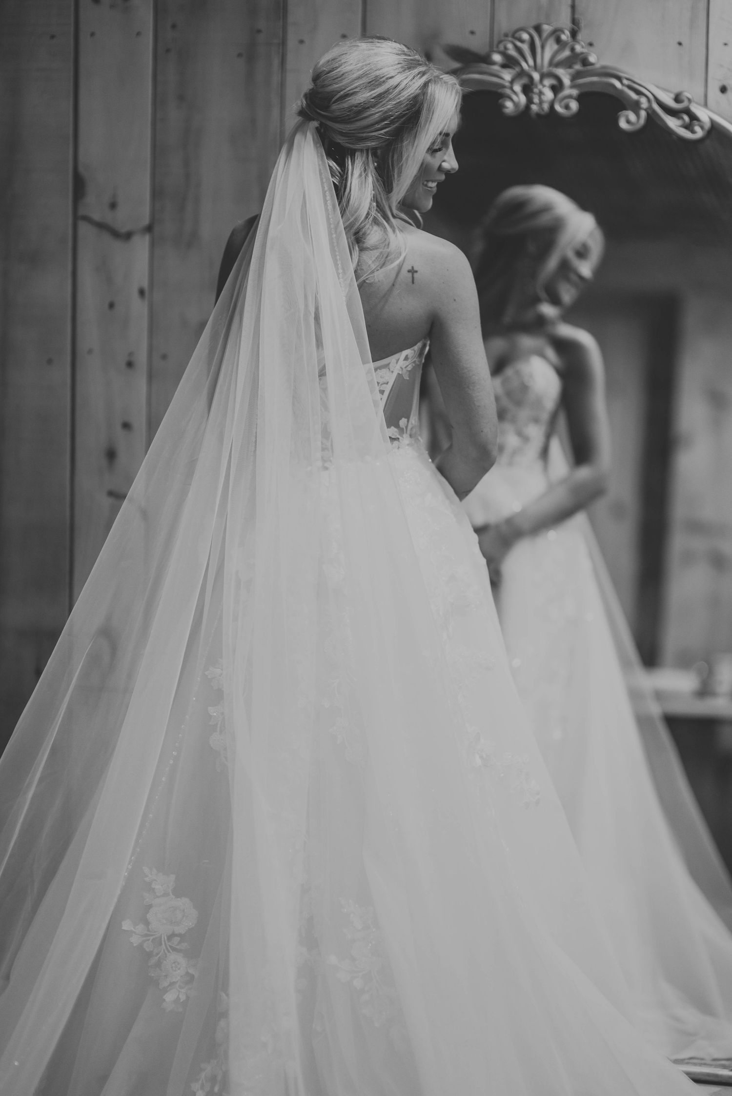 Black and white photo of a bride in a lace corset wedding dress with flowing veil looking in a mirror.