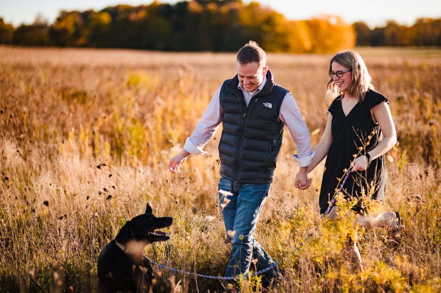 A couple walks with their black dog through a golden autumn field at sunset.