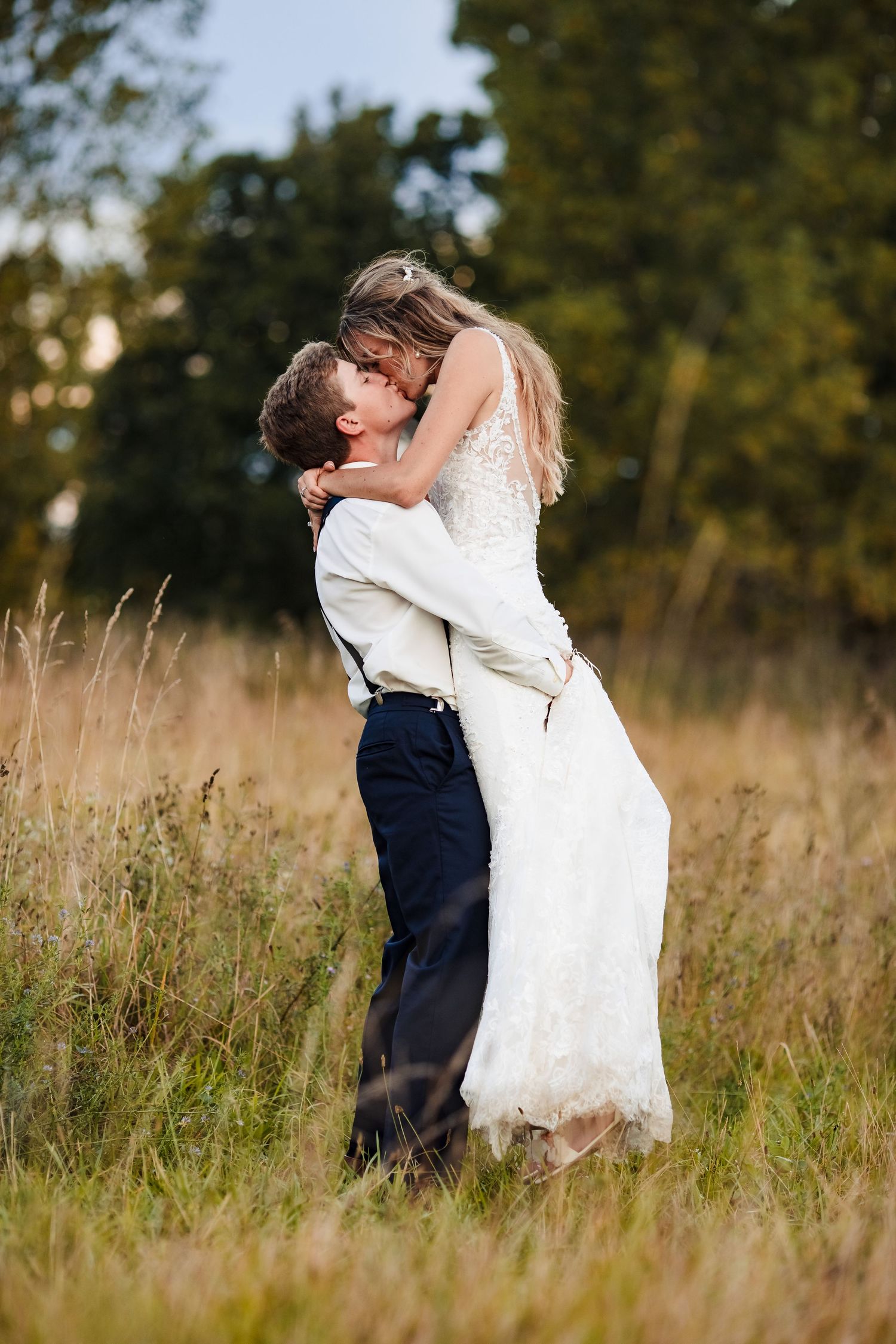 A romantic wedding couple embraces in a golden field at sunset with trees in the background.
