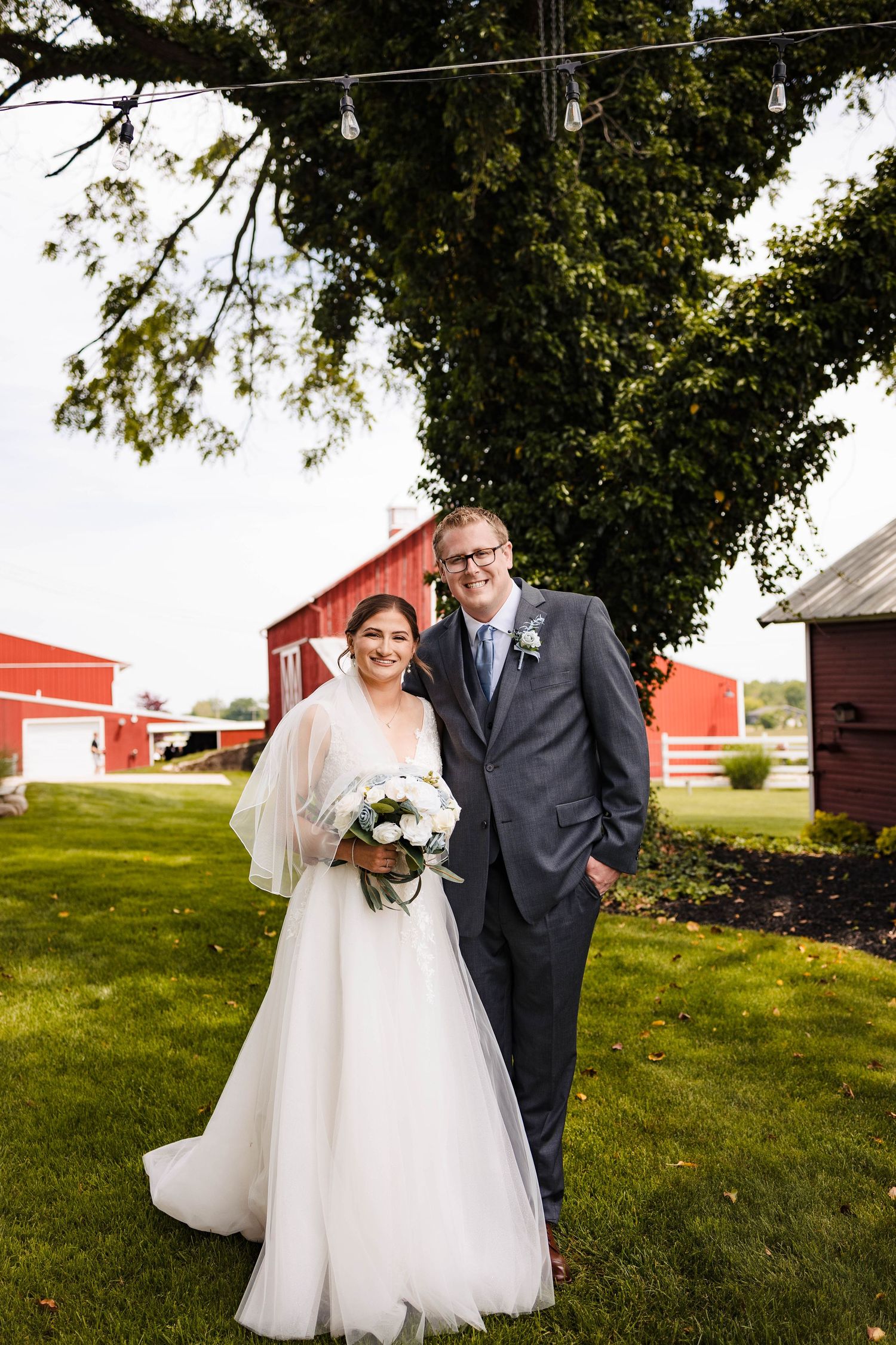 A bride and groom pose together on a grassy lawn with red barns in the background during a rustic farm wedding.