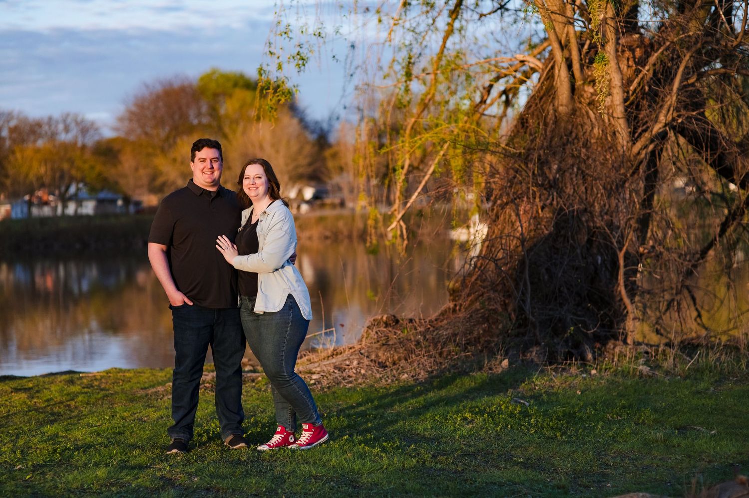 A couple poses together by a tranquil lake during golden hour with autumn trees in the background.